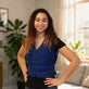 A smiling young woman with long curly brown hair stands indoors wearing a royal blue adjustable cooling zippered vest over a black short-sleeve top. The photo is taken in a softly lit room with plants and a window in the background.