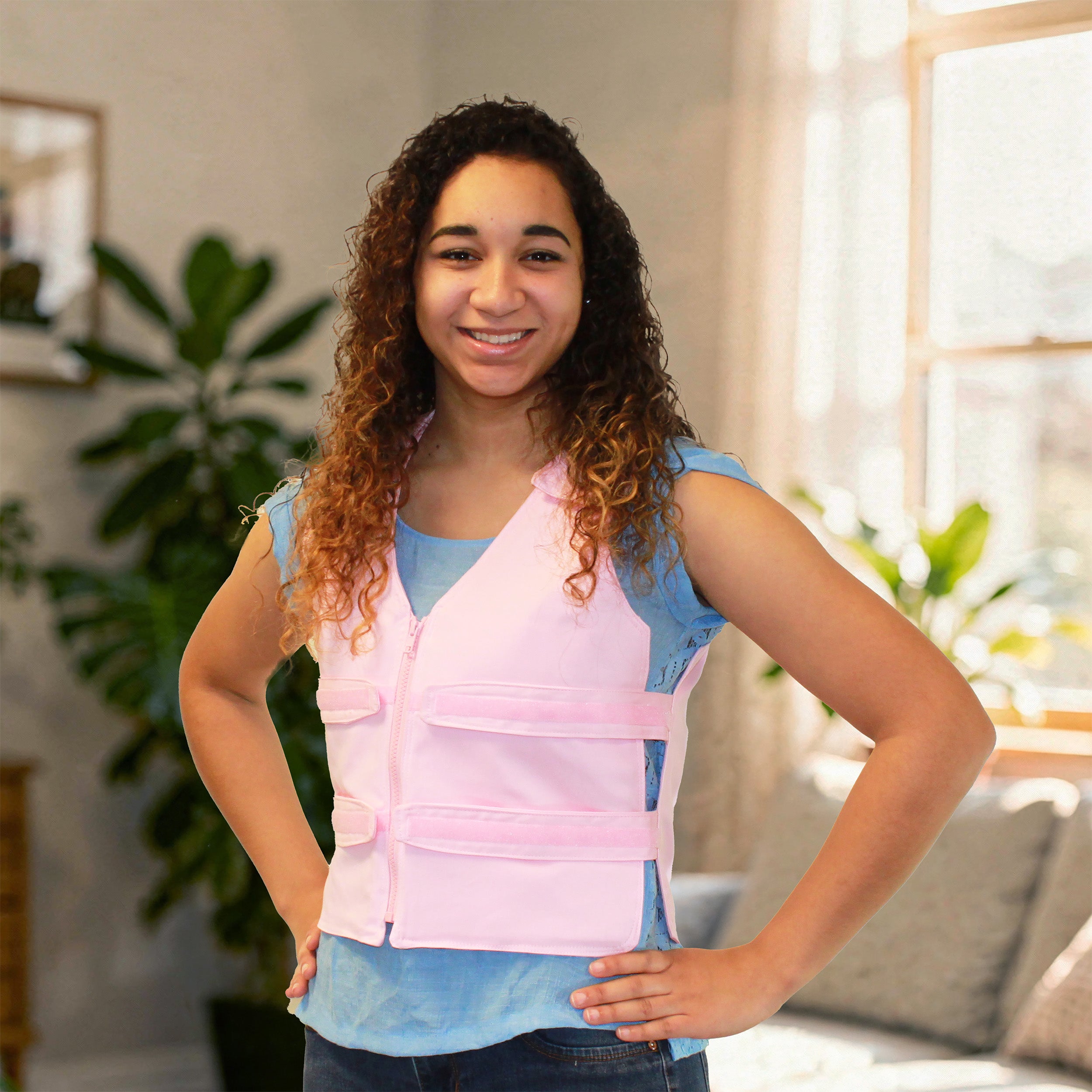 A smiling woman with curly brown hair wearing a light pink adjustable cooling vest over a light blue t-shirt, standing in a brightly lit room with plants and a window in the background.
