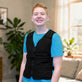 A smiling young man wearing a black adjustable cooling vest over a bright blue t-shirt, standing in a brightly lit living room with plants and a window in the background.