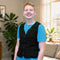 A smiling young man wearing a black adjustable cooling vest over a bright blue t-shirt, standing in a brightly lit living room with plants and a window in the background.