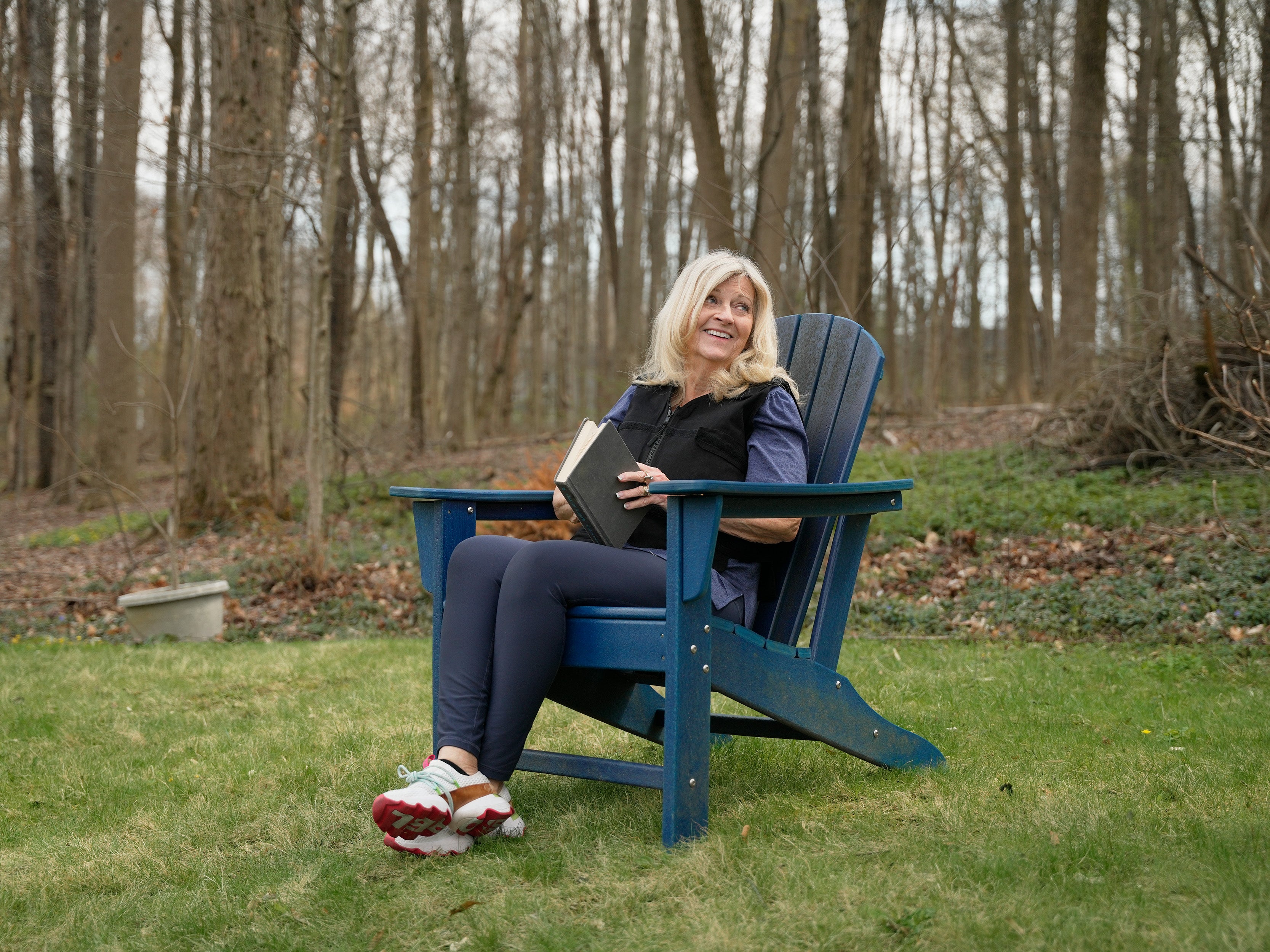 Woman sitting in a blue Adirondack chair outdoors in a wooded area reading a book wearing a black adjustable cooling vest over a blue top.