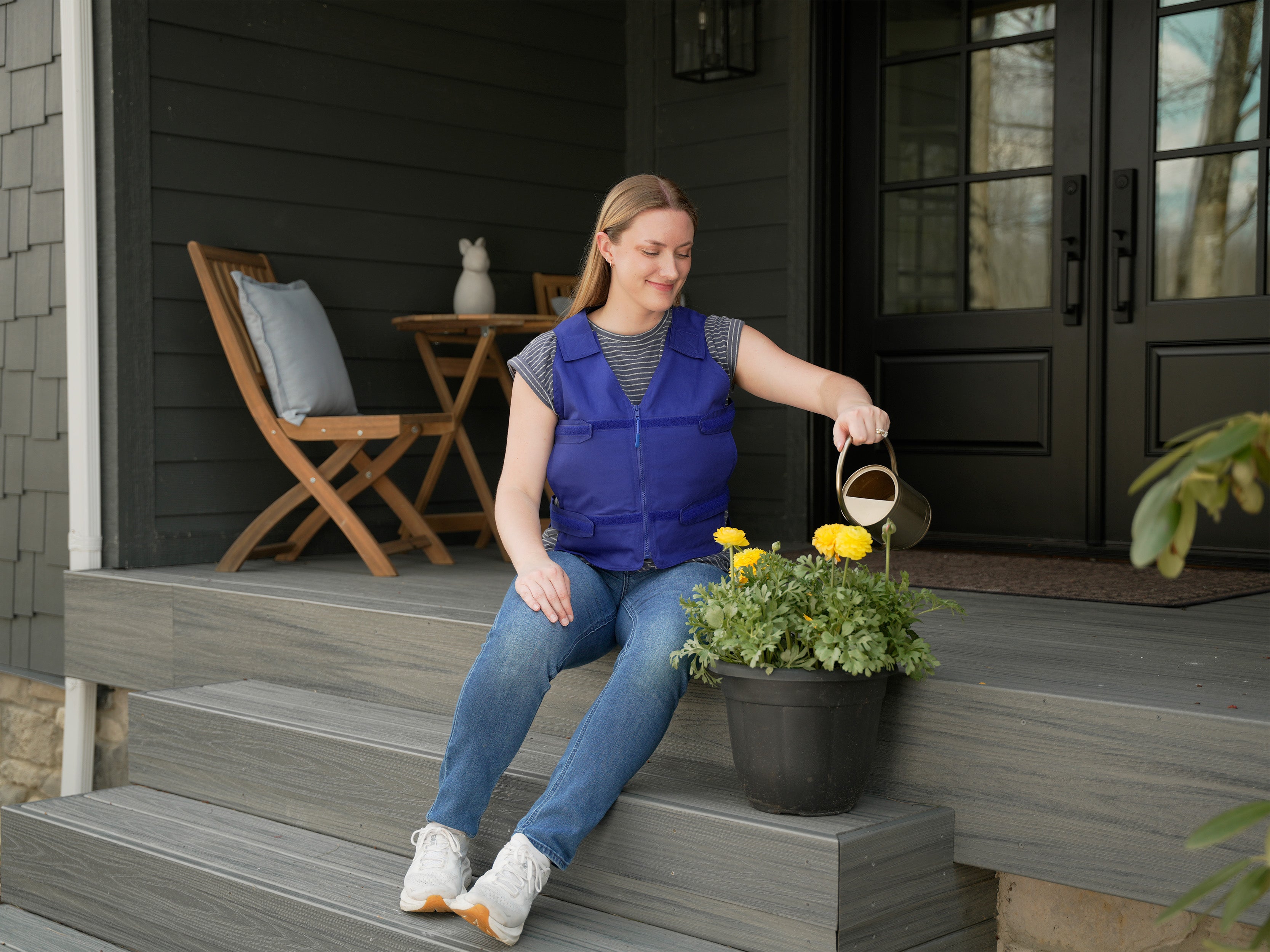 Woman sitting on a porch, watering plants with a small watering can wearing a blue adjustable cooling vest over a striped t-shirt.