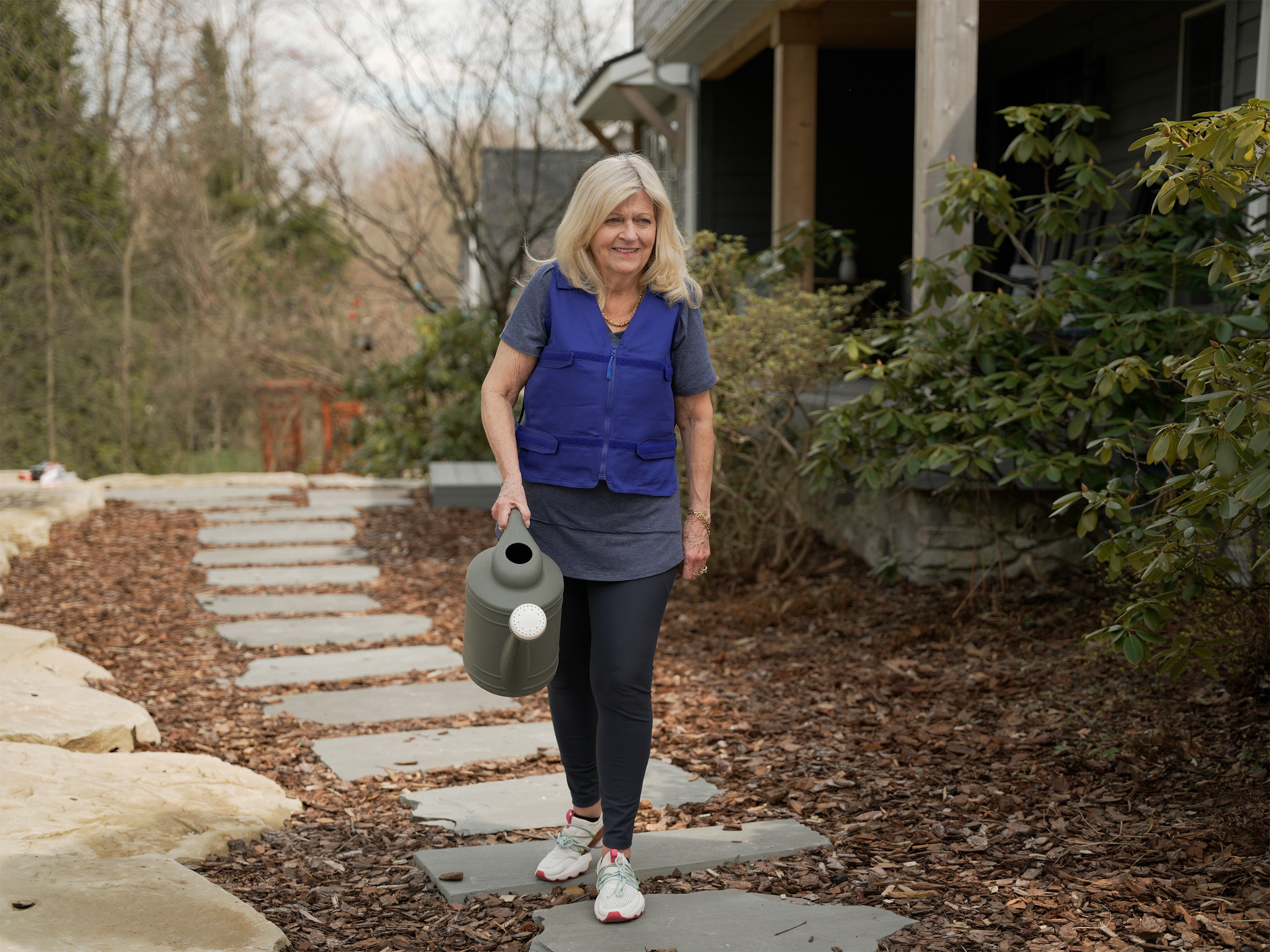 A woman walks along a stone path in a backyard, holding a gray watering can. She is wearing a blue Adjustable zipper cooling vest over a gray t-shirt, dark leggings, and sneakers.
