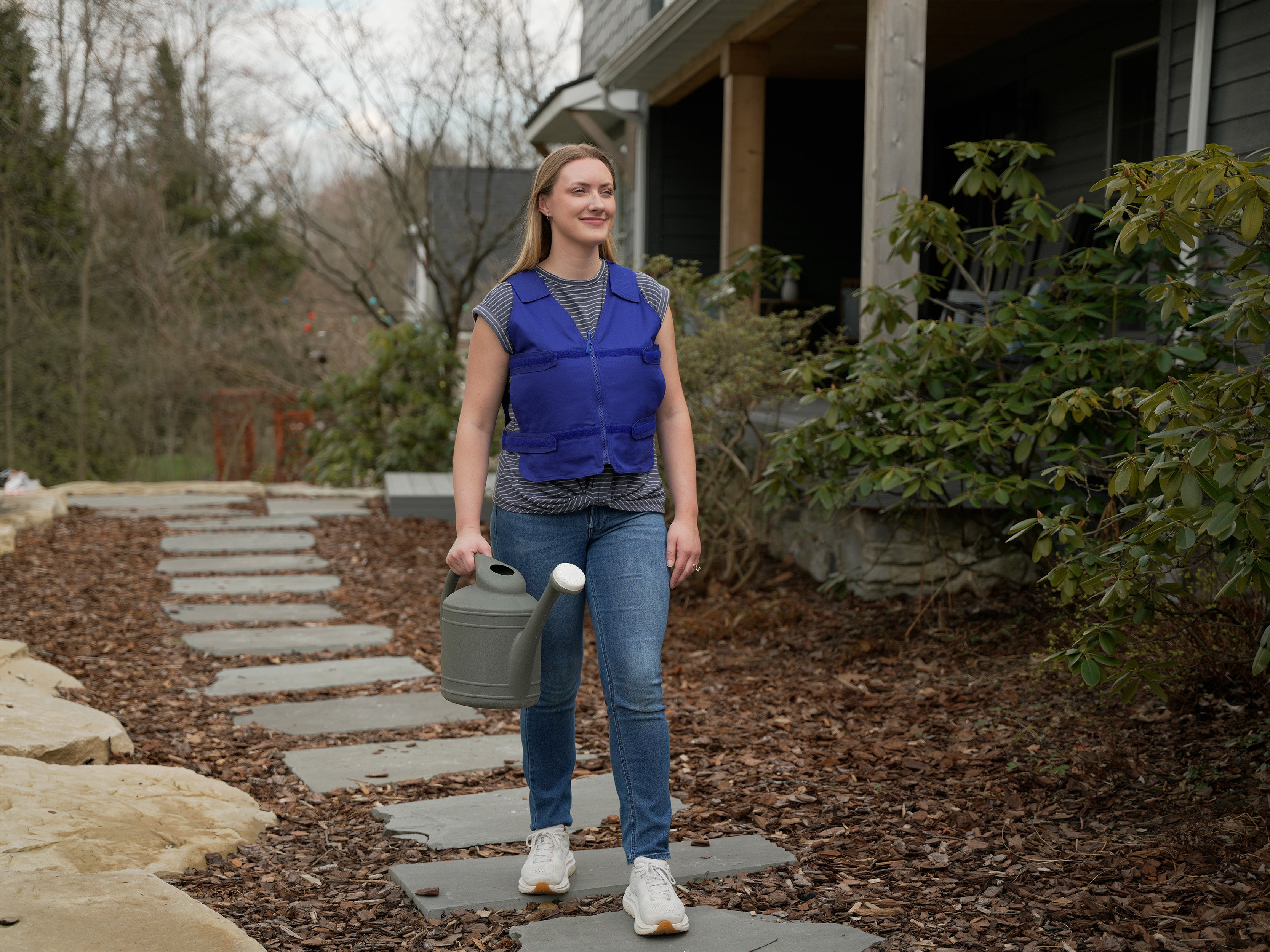 A woman walking on a stone path in front of a house, wearing a bright blue adjustable cooling vest over a gray shirt and blue jeans while holding a gray watering can.