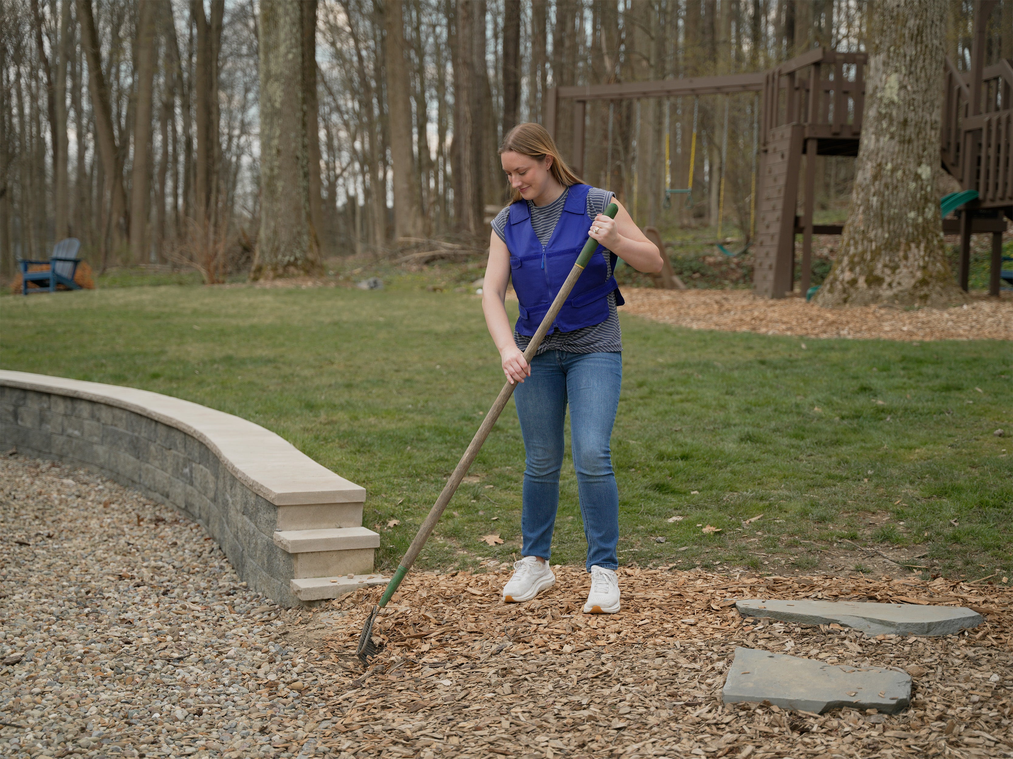 A woman wearing a blue adjustable cooling vest, gray t-shirt, and blue jeans is raking wood chips in an outdoor backyard setting with trees and a wooden playset in the background.