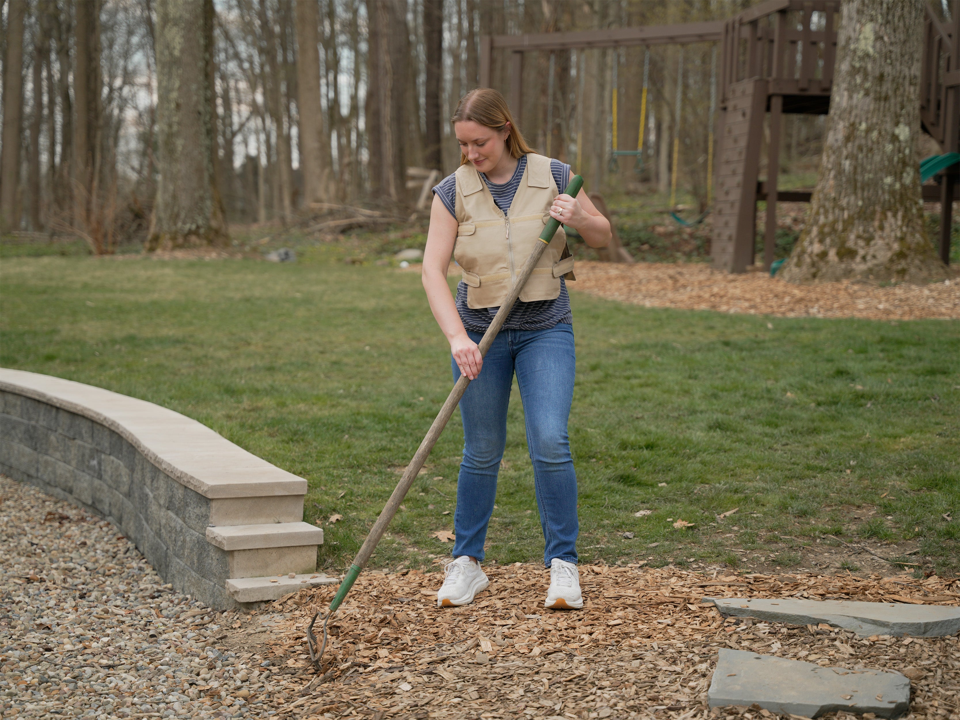 A woman in a beige adjustable cooling vest and blue jeans is raking mulch in a backyard with trees and a wooden playset in the background.