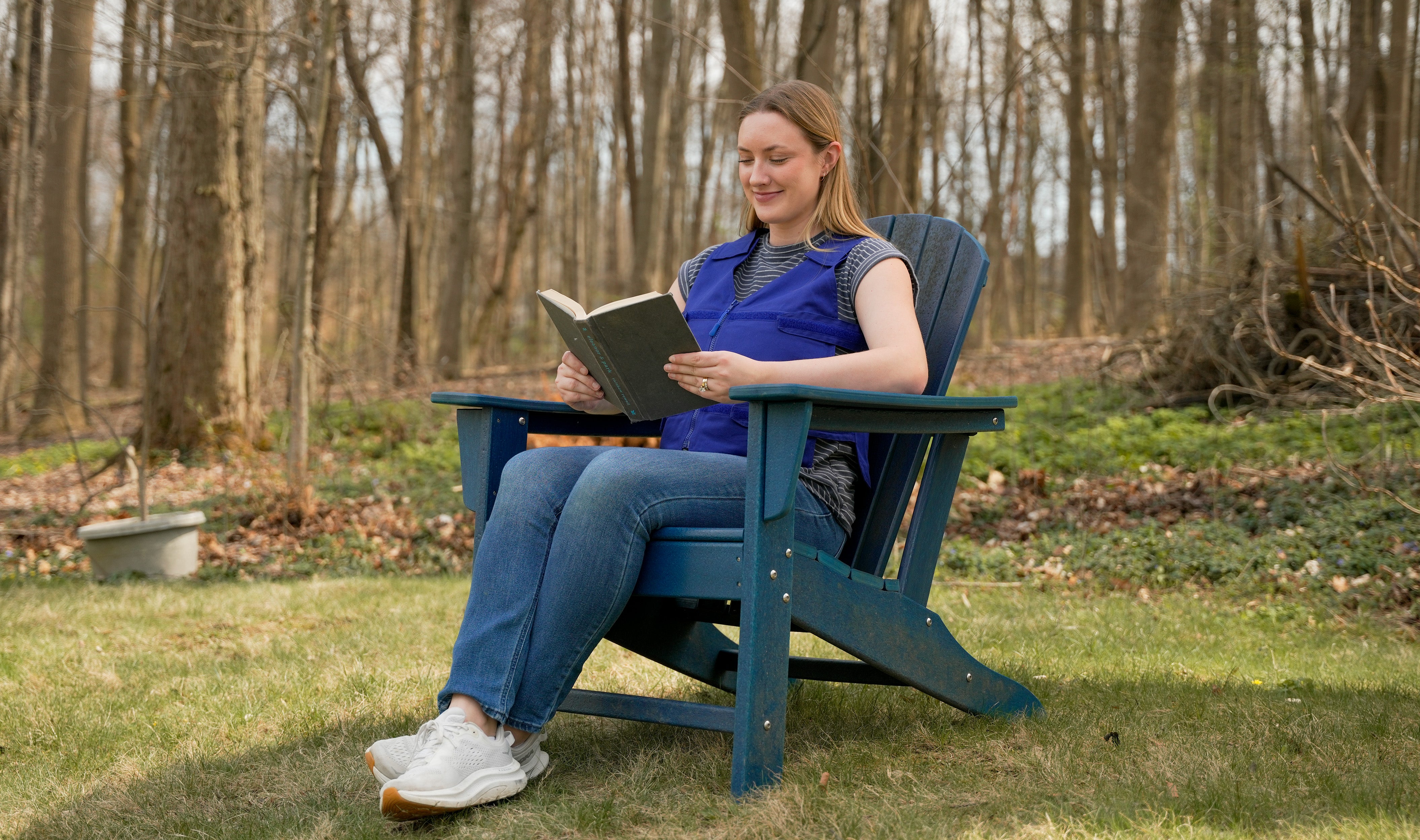A woman sits in a blue wooden Adirondack chair reading a book and wearing a blue adjustable cooling vest in a grassy backyard with a wooded background.