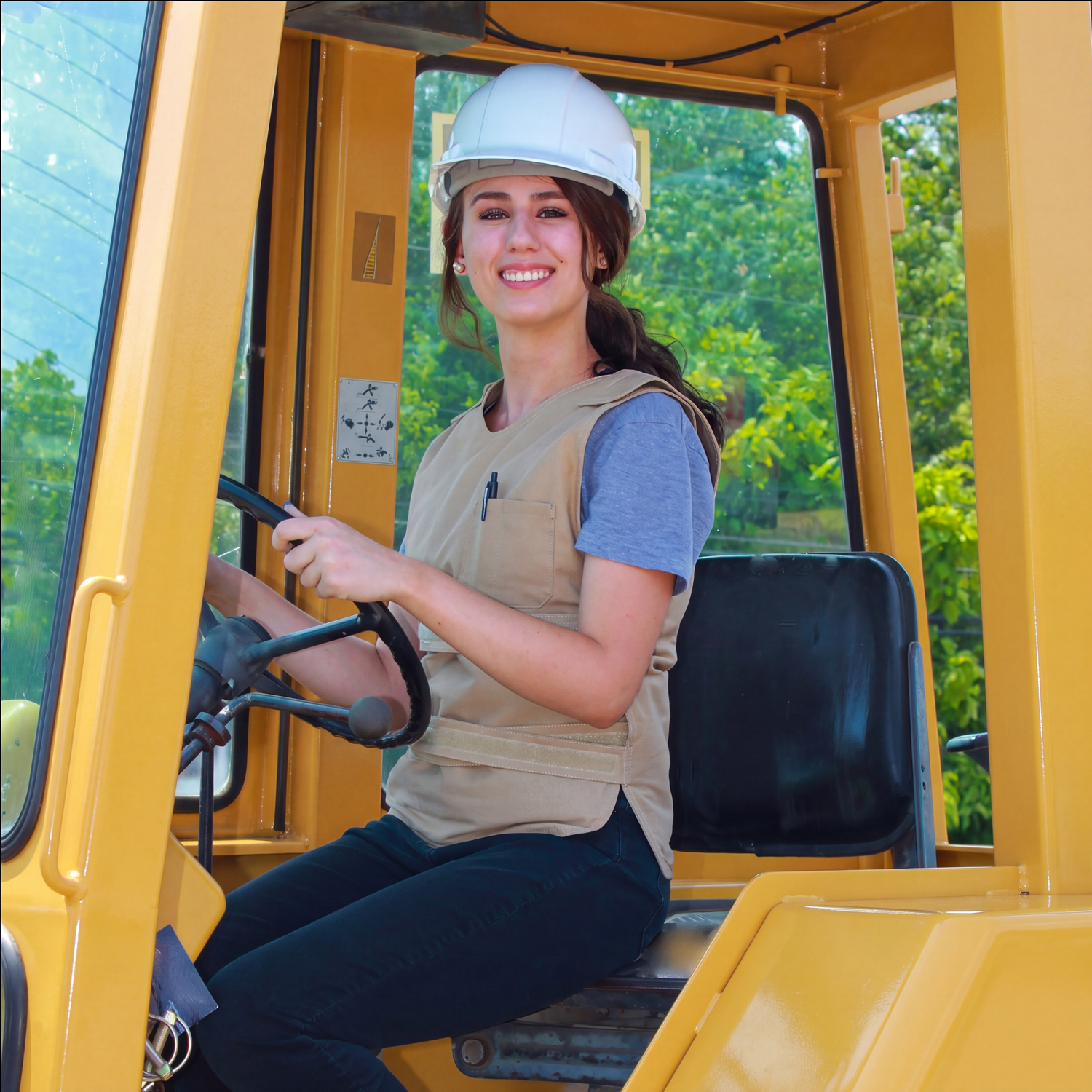 A smiling woman wearing a white hard hat and a beige adjustable cooling poncho vest over a blue t-shirt sits inside the cab of a yellow construction vehicle, holding the steering wheel.