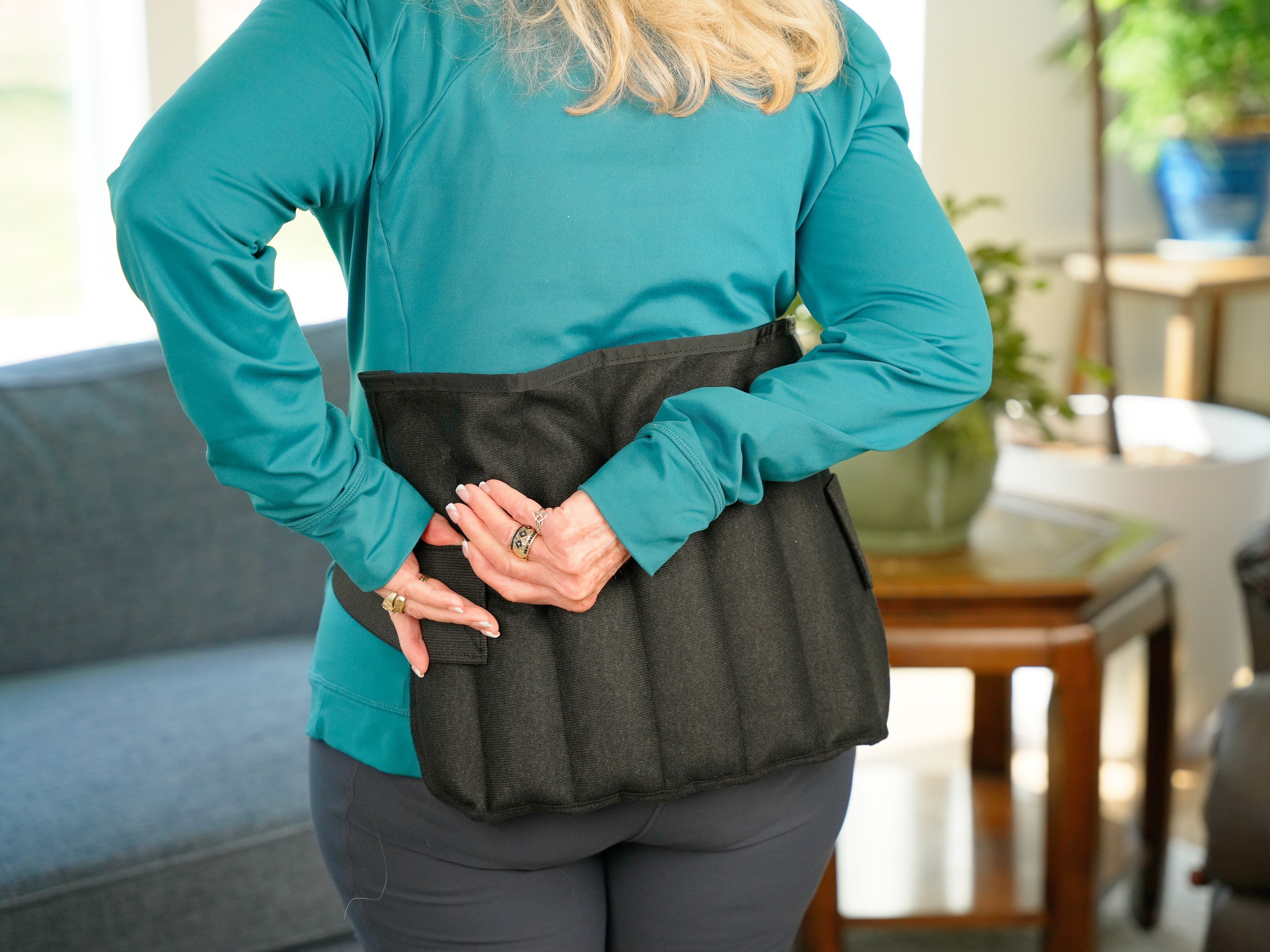 A woman wearing a teal long-sleeve shirt and dark gray leggings is shown from the back, wearing a black Moist Heat Thera-Temp Wrap around her lower back. The setting appears to be a blurred indoor living room environment.