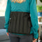 A woman wearing a teal long-sleeve shirt and dark gray leggings is shown from the back, wearing a black Moist Heat Thera-Temp Wrap around her lower back. The setting appears to be a blurred indoor living room environment.