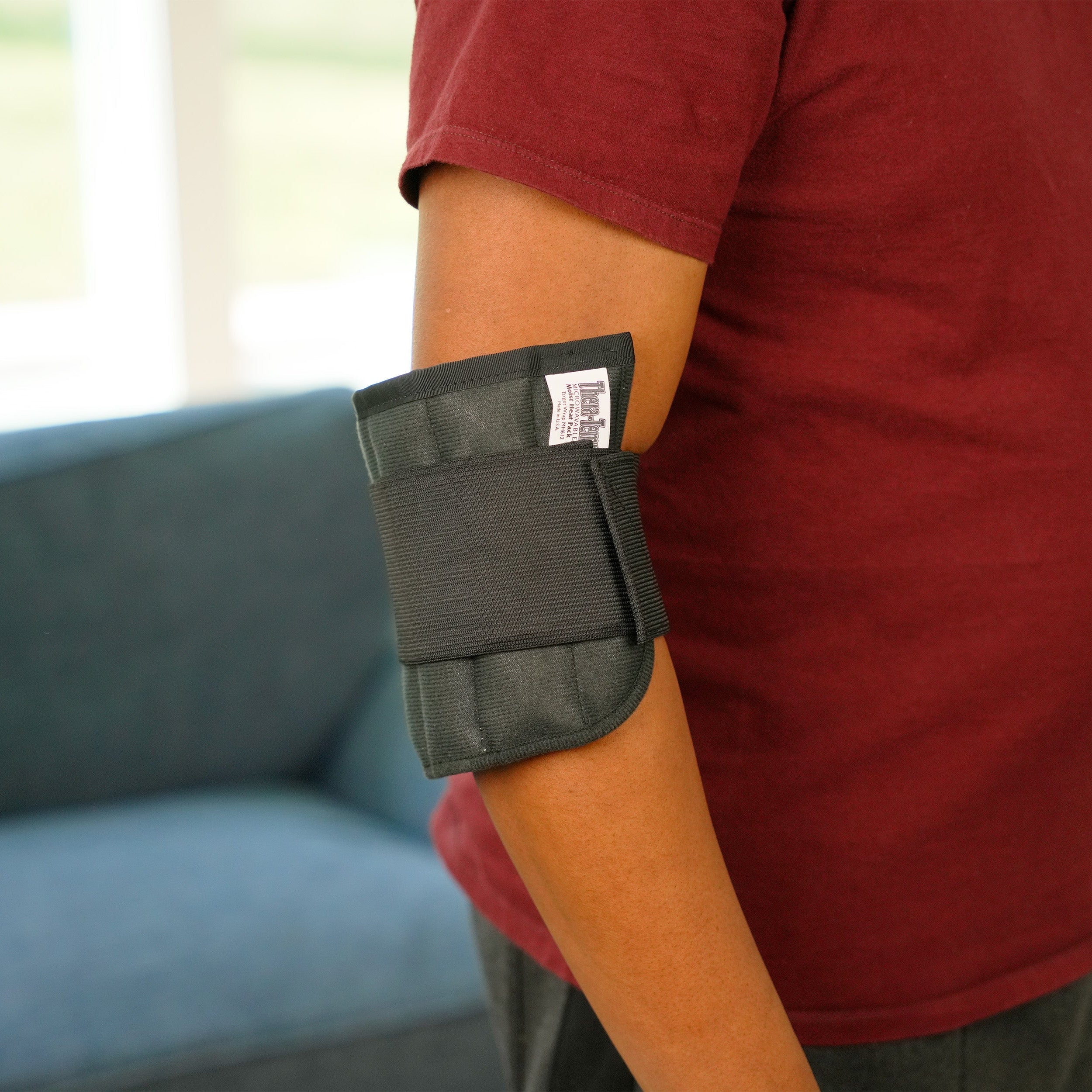 A person wearing a Moist Heat Thera-Temp Wrap on his elbow, shown from the side in an indoor setting with a blue sofa in the background.