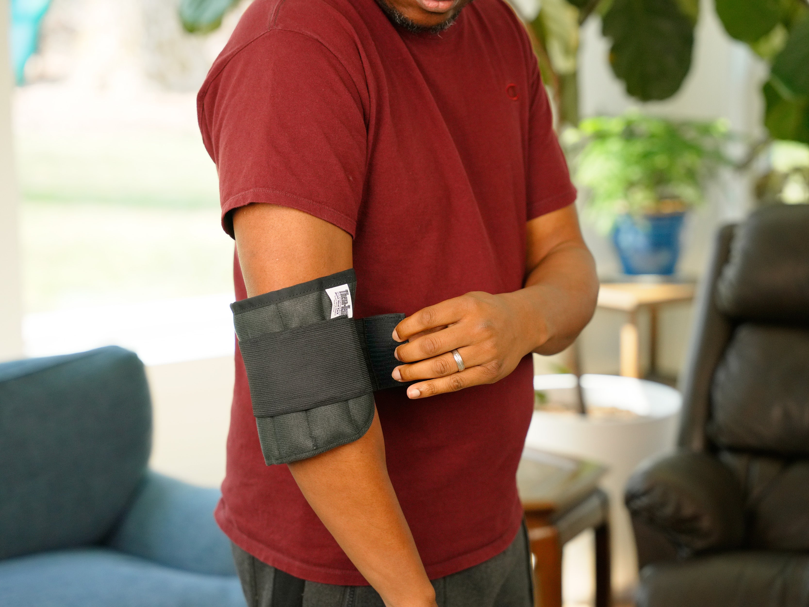 A person wearing a Moist Heat Thera-Temp Wrap on his elbow, shown from the side in an indoor setting with a blue sofa in the background.