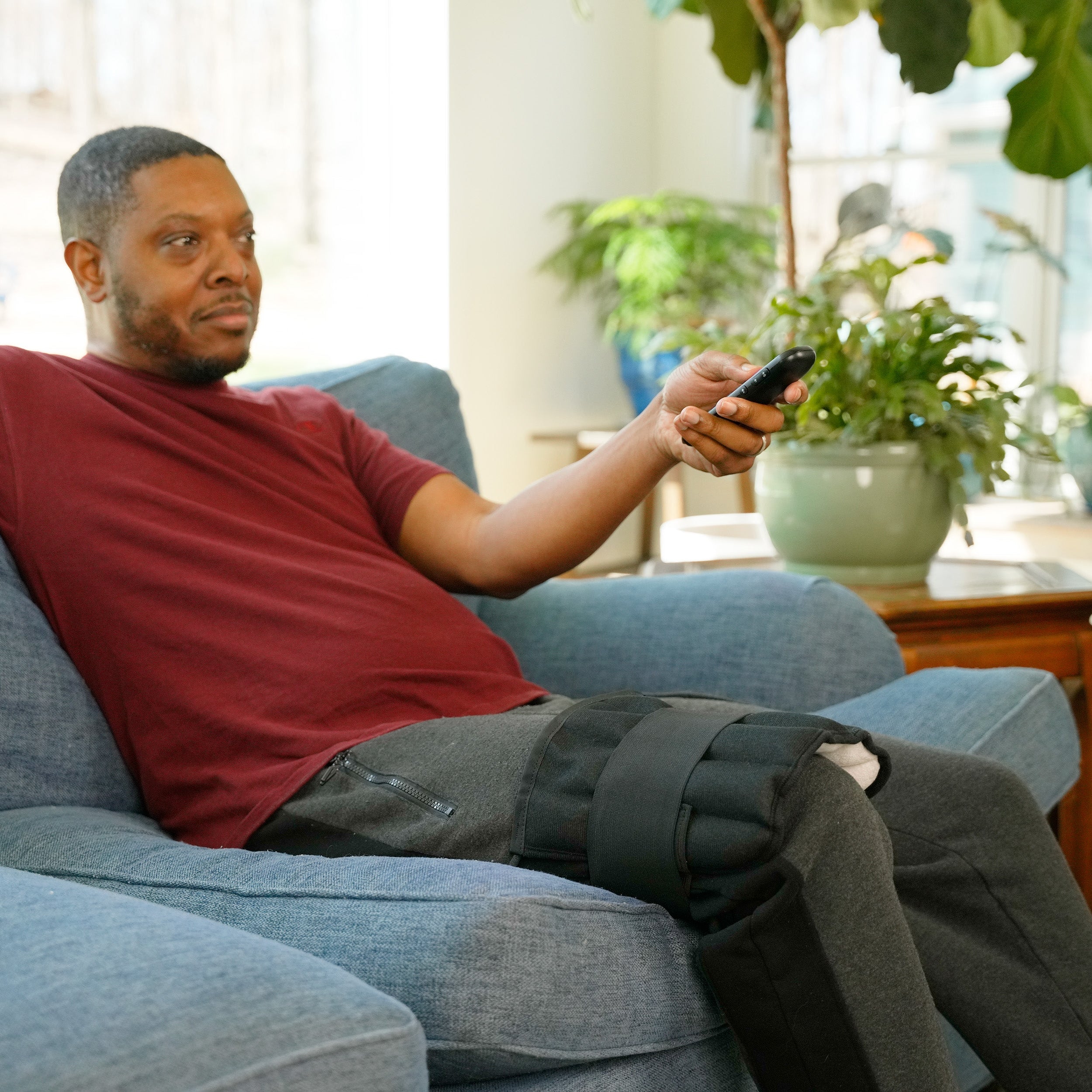 A man reclining on a blue couch in a living room, holding a remote control. He is wearing a black leg Moist Heat Thera-Temp Wrap on his right leg.