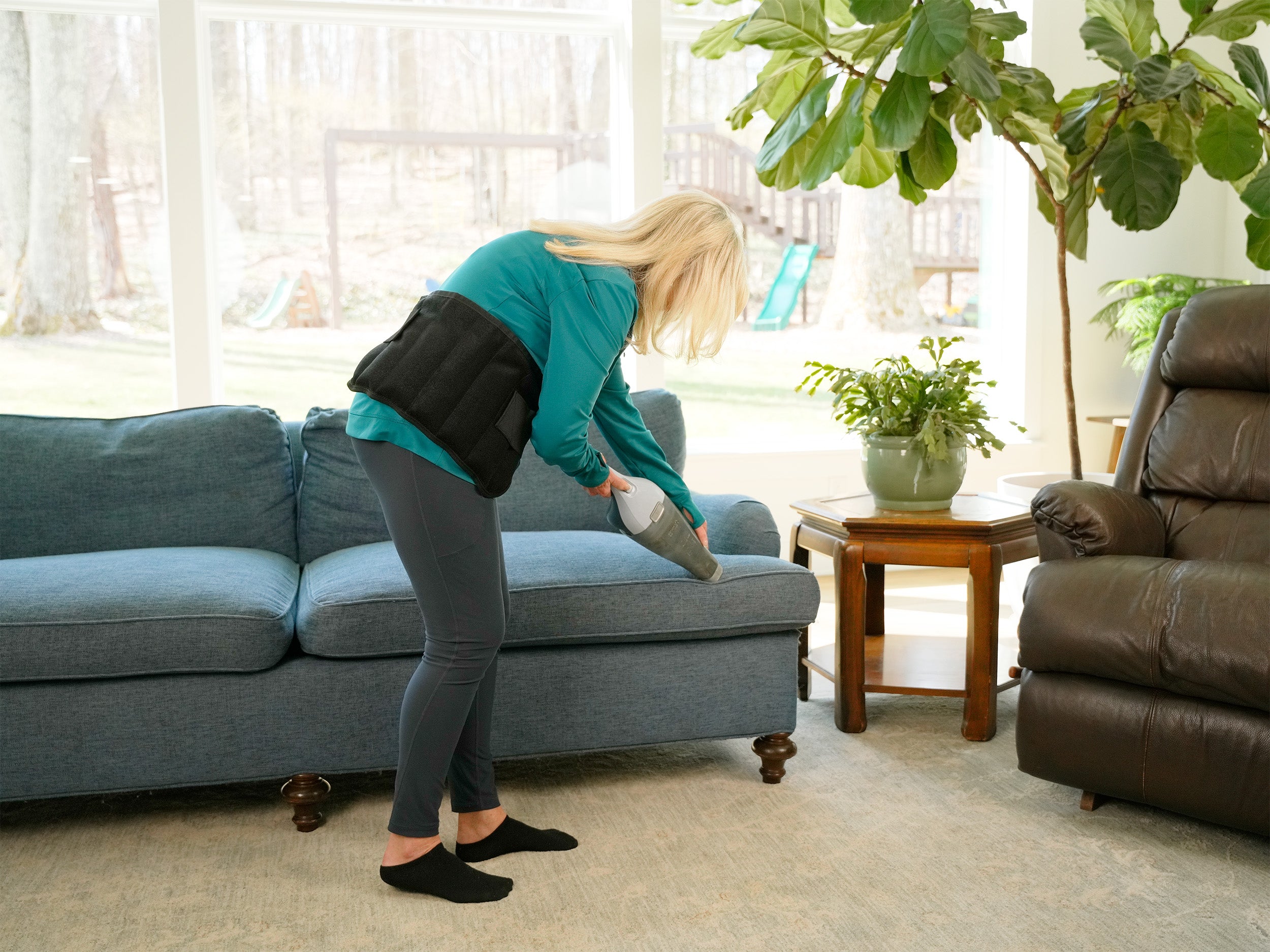 A woman in a teal top and gray leggings wearing a Moist Heat Thera-Temp Wrap around her waist. She is using a gray handheld vacuum cleaner on a blue fabric sofa in a bright living room with a brown recliner, potted plants, and large windows.