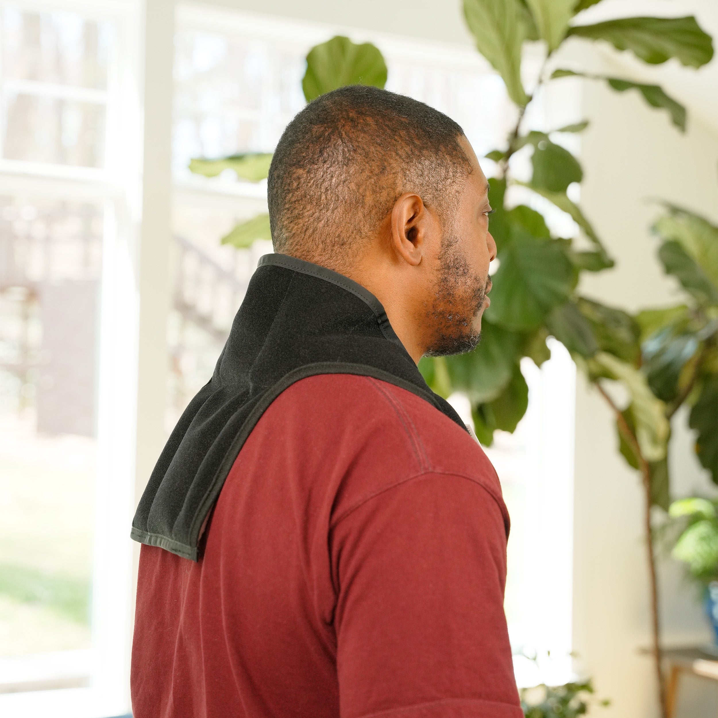 A man in a red t-shirt shown from the back and side profile, wearing a Moist Heat Thera-Temp Wrap over his shoulders in a brightly lit indoor room with a large green plant in the background.
