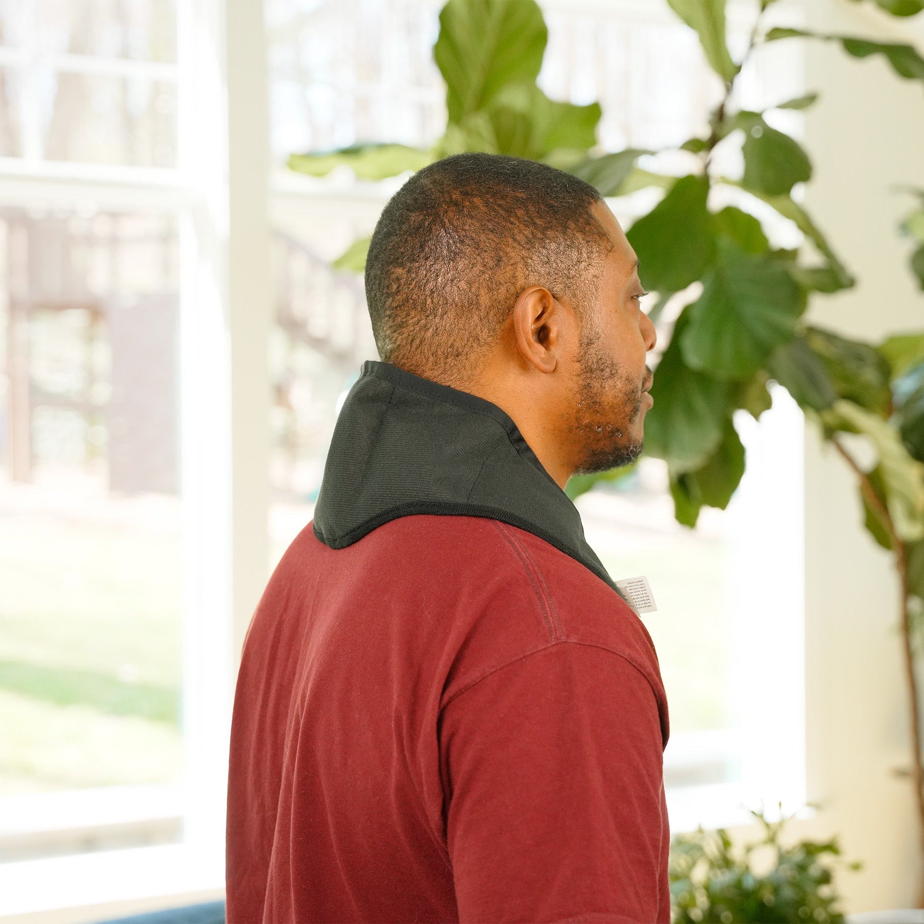 A man shown from the back and side profile wearing a red long-sleeve shirt wearing a Moist Heat Thera-Temp wrap around his neck, standing in a brightly lit room with large green houseplants near a window.