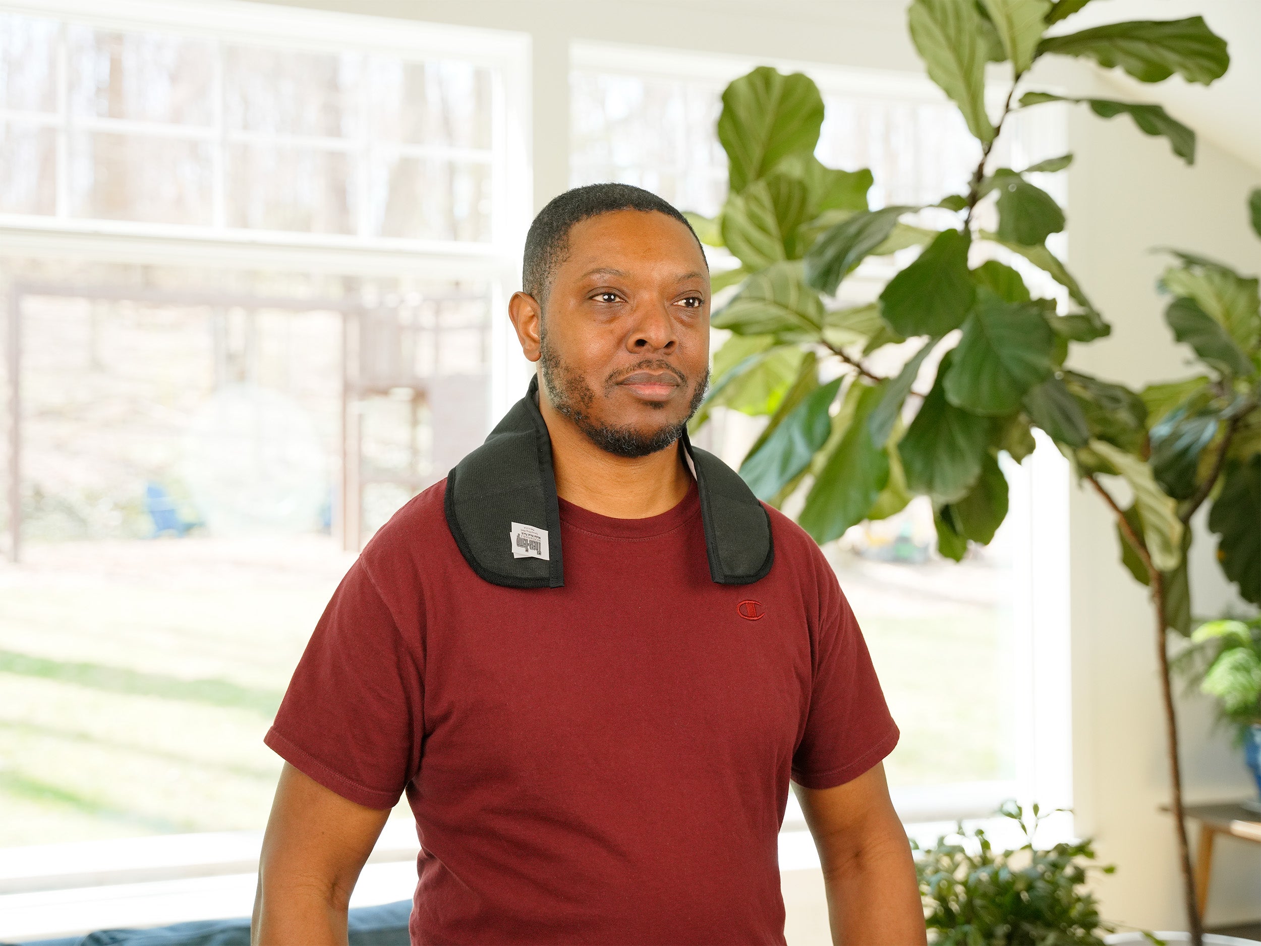 A man wearing a Moist Heat Thera-Temp wrap around his neck over a maroon t-shirt, standing indoors in front of a bright window and a large green fiddle-leaf fig plant.