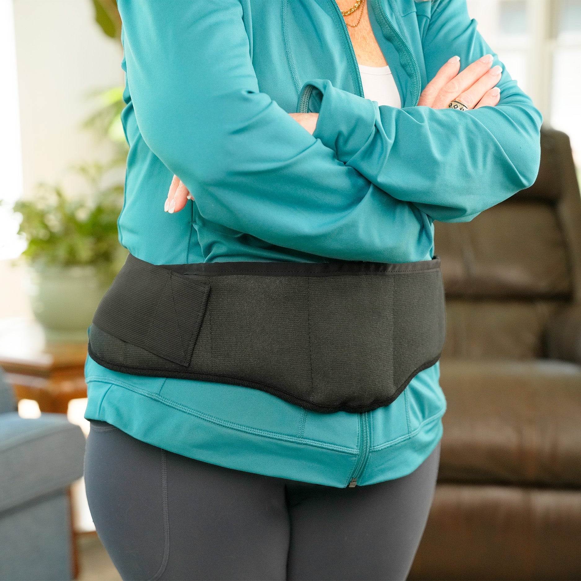 A person wearing a teal athletic zip-up jacket and gray leggings stands with arms crossed, wearing a Moist Heat Thera-Temp Wrap around their midsection in a living room setting.
