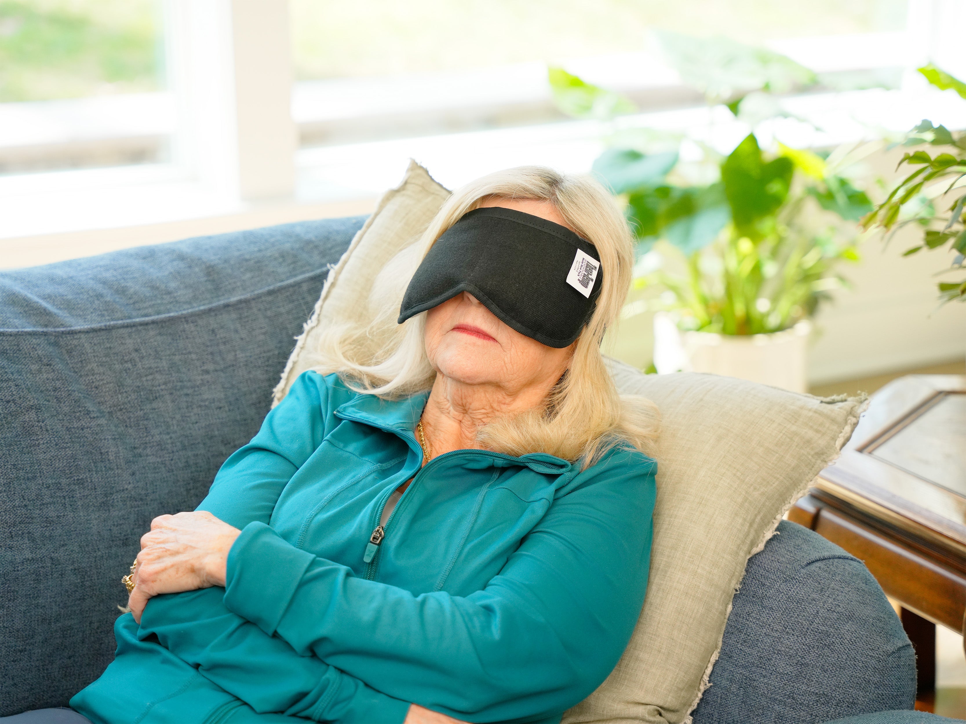 A woman wearing a black Moist Heat Thera-Temp Eye & Sinus mask and a teal zip-up jacket stands indoors in a bright living room with large green plants and windows in the background.