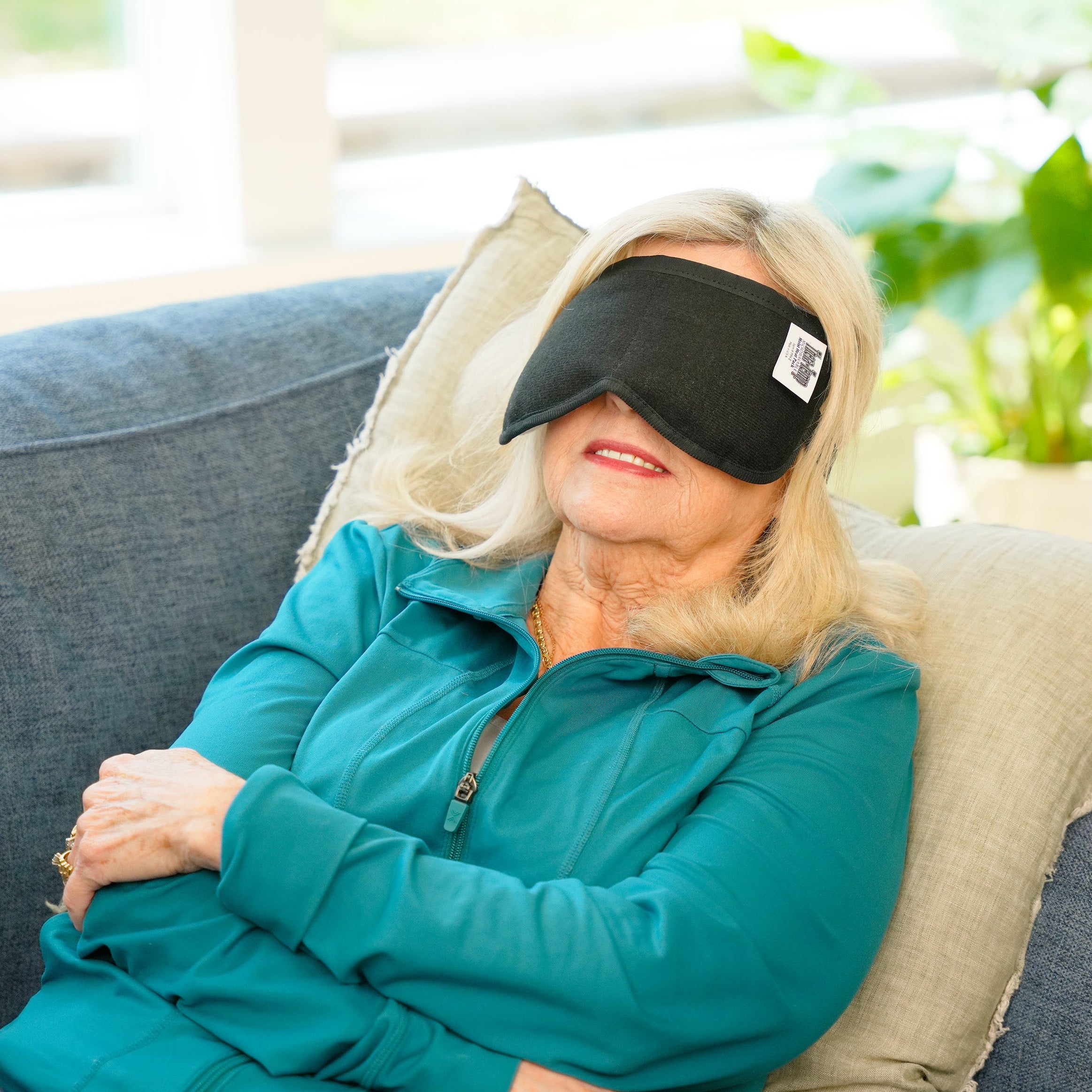 Woman in living room wearing a thera-temp® moist heat eye wrap over her eyes and nose while laying on a blue coach with windows and plants behind her