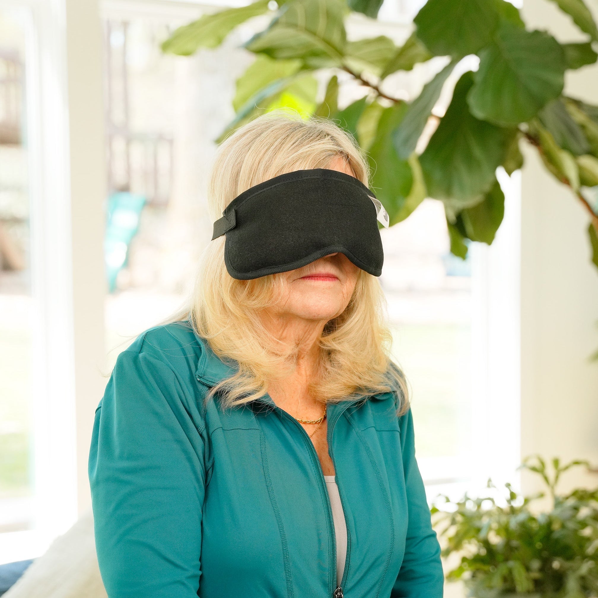 A woman wearing a black Moist Heat Thera-Temp Eye & Sinus mask and a teal zip-up jacket stands indoors in a bright living room with large green plants and windows in the background.