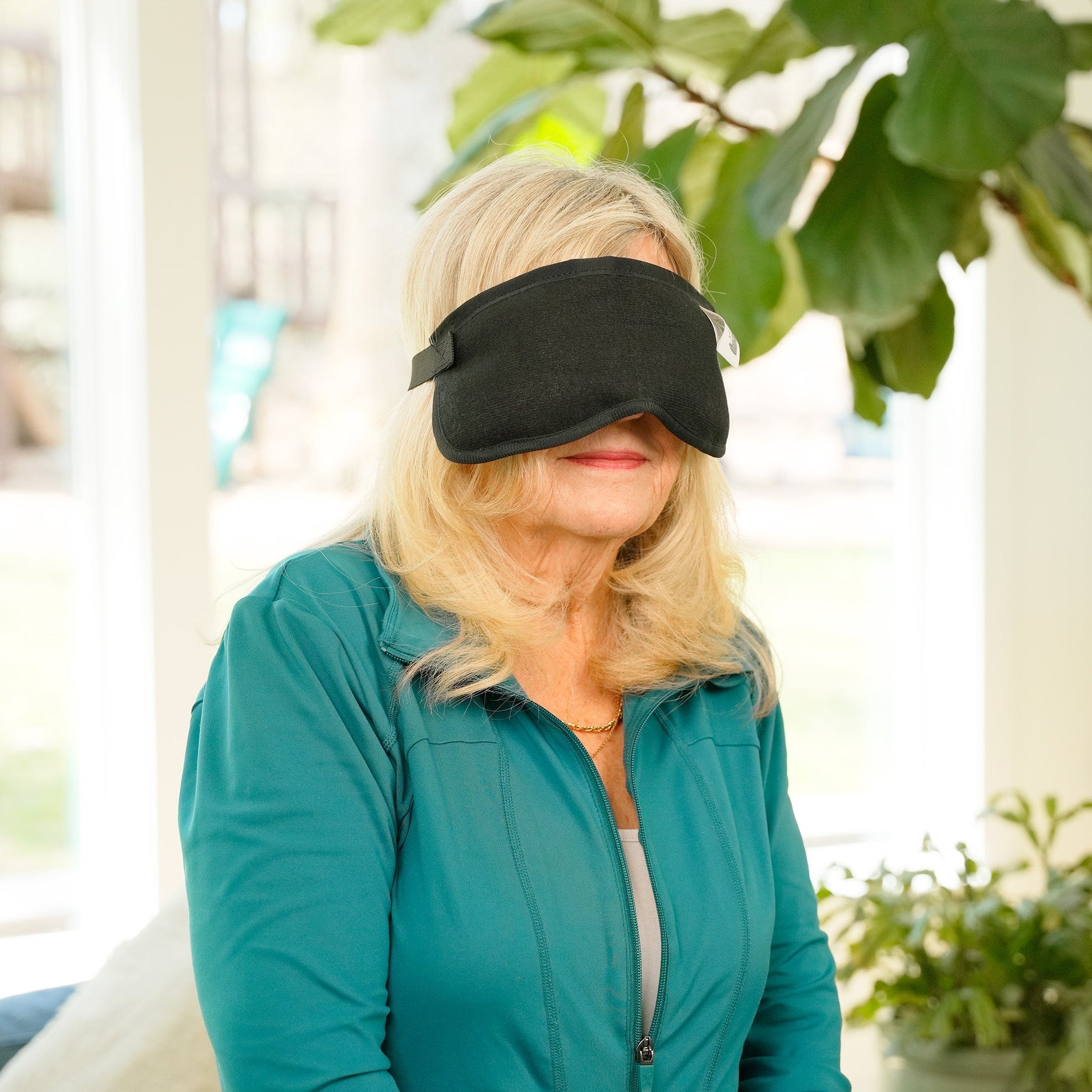 Woman in living room wearing a thera-temp® moist heat eye wrap over her eyes and nose with windows and plants behind her