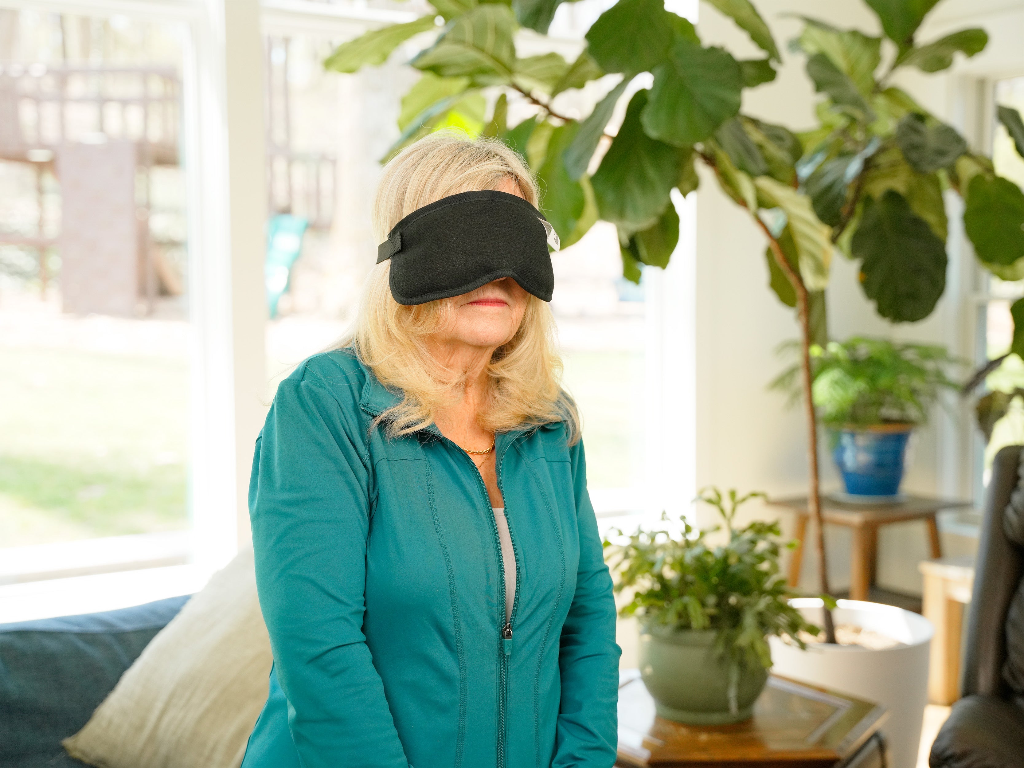 A woman wearing a black Moist Heat Thera-Temp Eye & Sinus mask and a teal zip-up jacket stands indoors in a bright living room with large green plants and windows in the background.