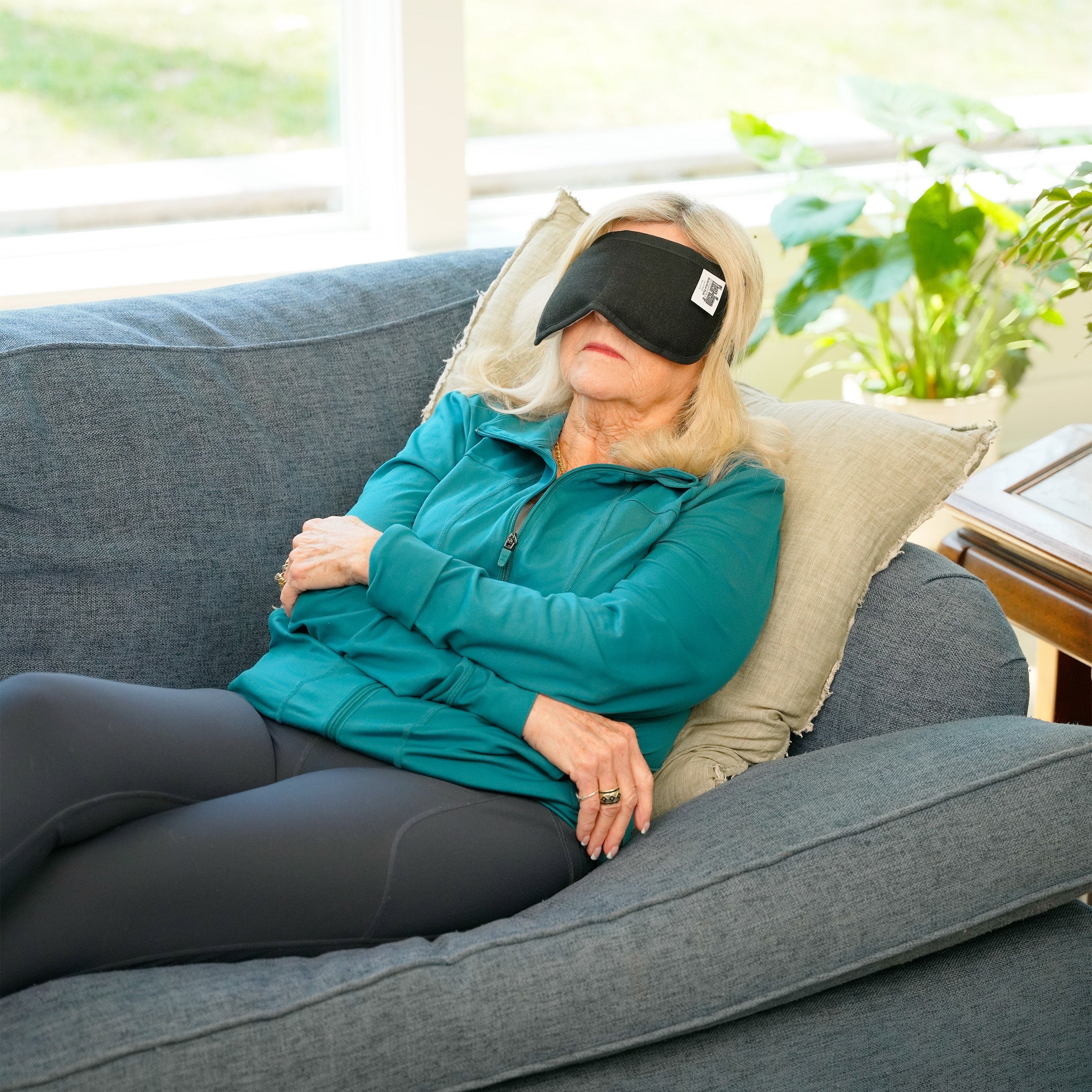 A woman wearing a black Moist Heat Thera-Temp Eye & Sinus mask and a teal zip-up jacket stands indoors in a bright living room with large green plants and windows in the background.