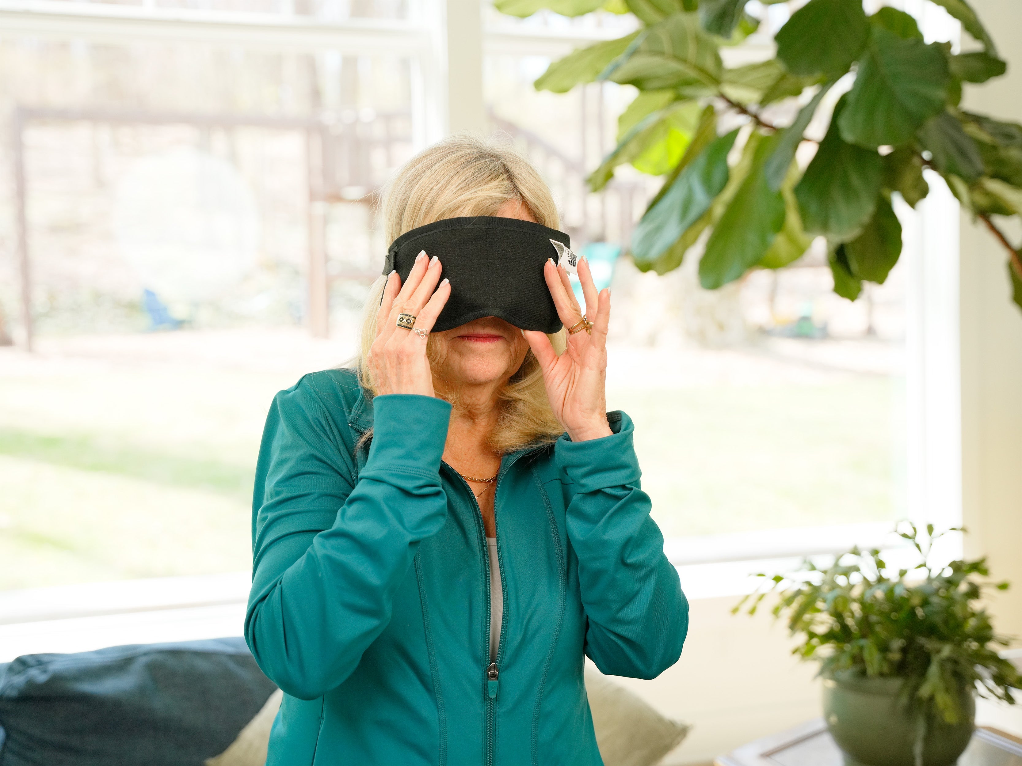 A woman wearing a black Moist Heat Thera-Temp Eye & Sinus mask and a teal zip-up jacket stands indoors in a bright living room with large green plants and windows in the background.