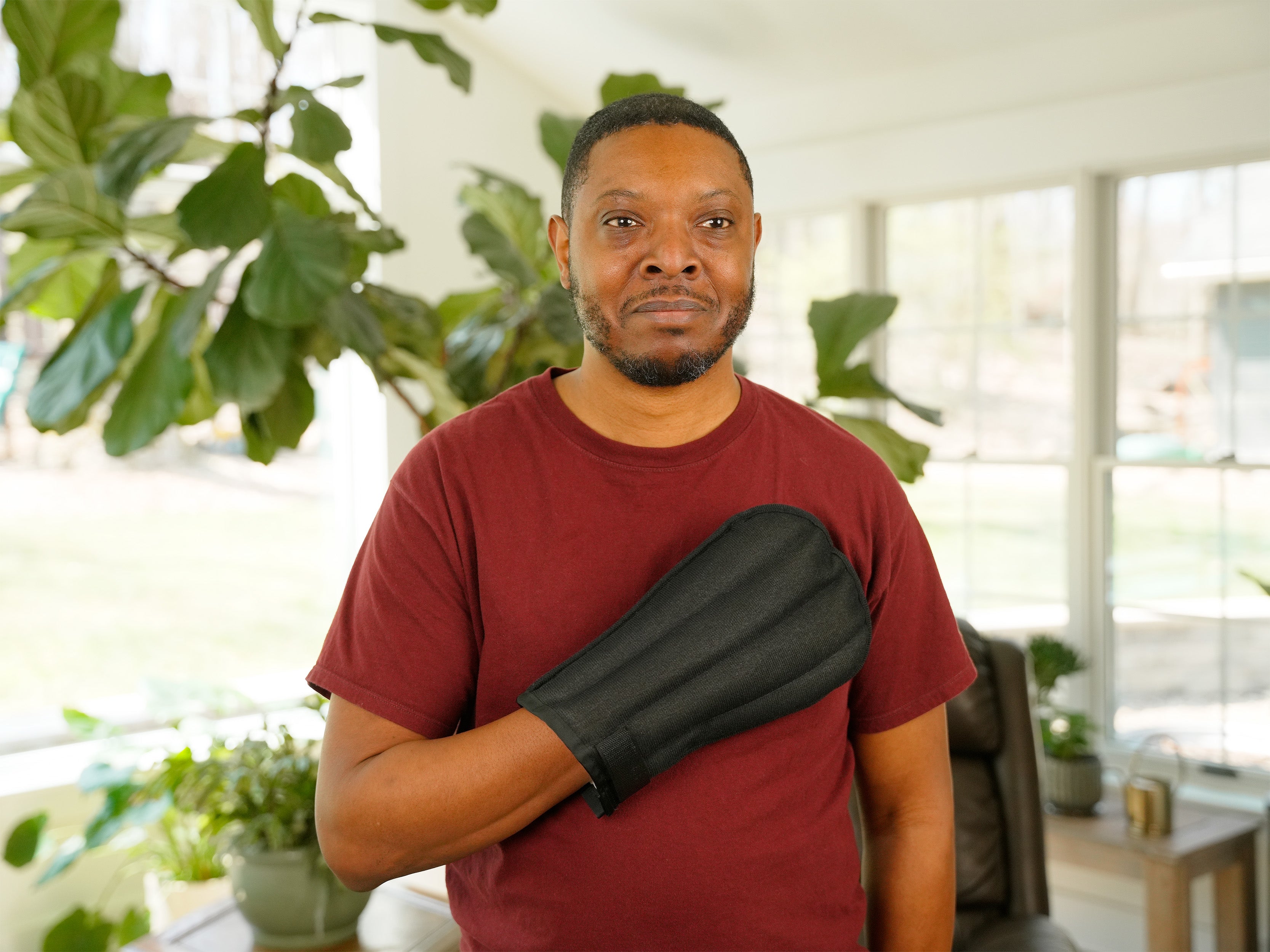A man wearing a Moist Heat Thera-Temp Mitt on his hand, paired with a maroon t-shirt, shown in a softly blurred indoor setting.
