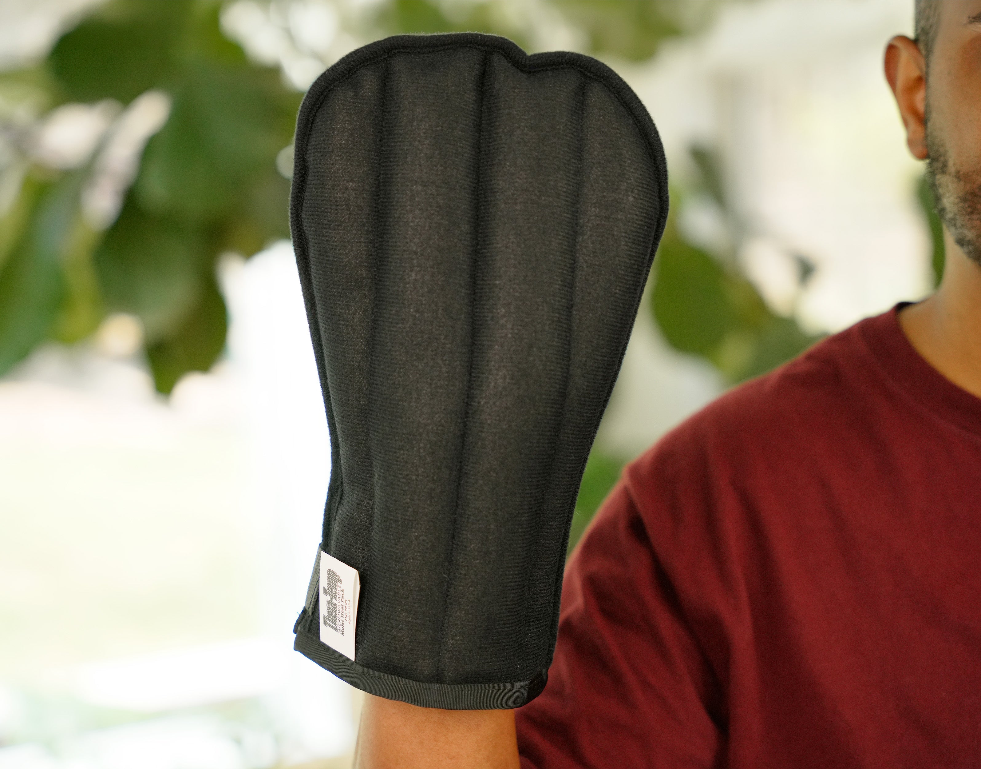 A man wearing a Moist Heat Thera-Temp Mitt on his hand, paired with a maroon t-shirt, shown in a softly blurred indoor setting.