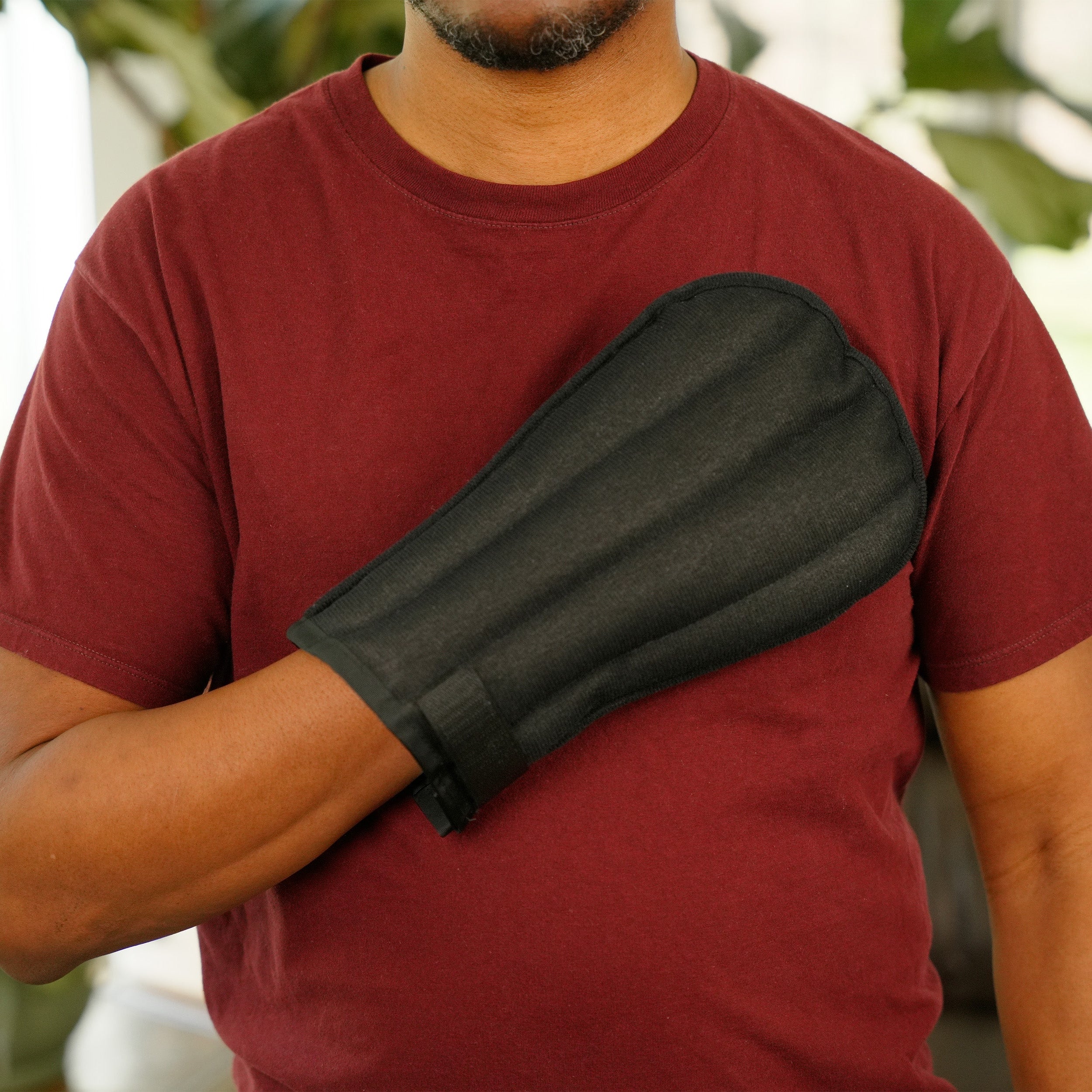A man wearing a Moist Heat Thera-Temp Mitt on his hand, paired with a maroon t-shirt, shown in a softly blurred indoor setting.