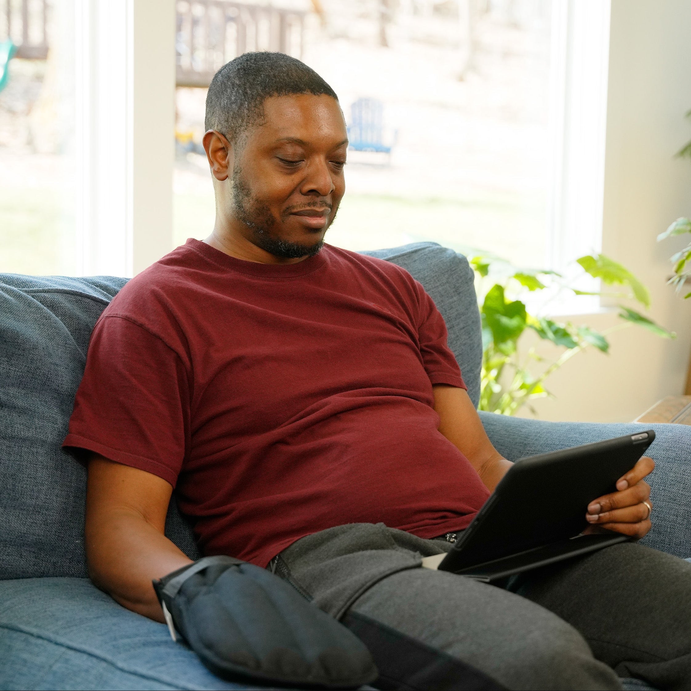 A man sitting on a blue couch in a bright living room, holding a black tablet. He is wearing Moist Heat Thera-Temp Mitt and  a maroon t-shirt and gray sweatpants, with house plants and a window in the background.