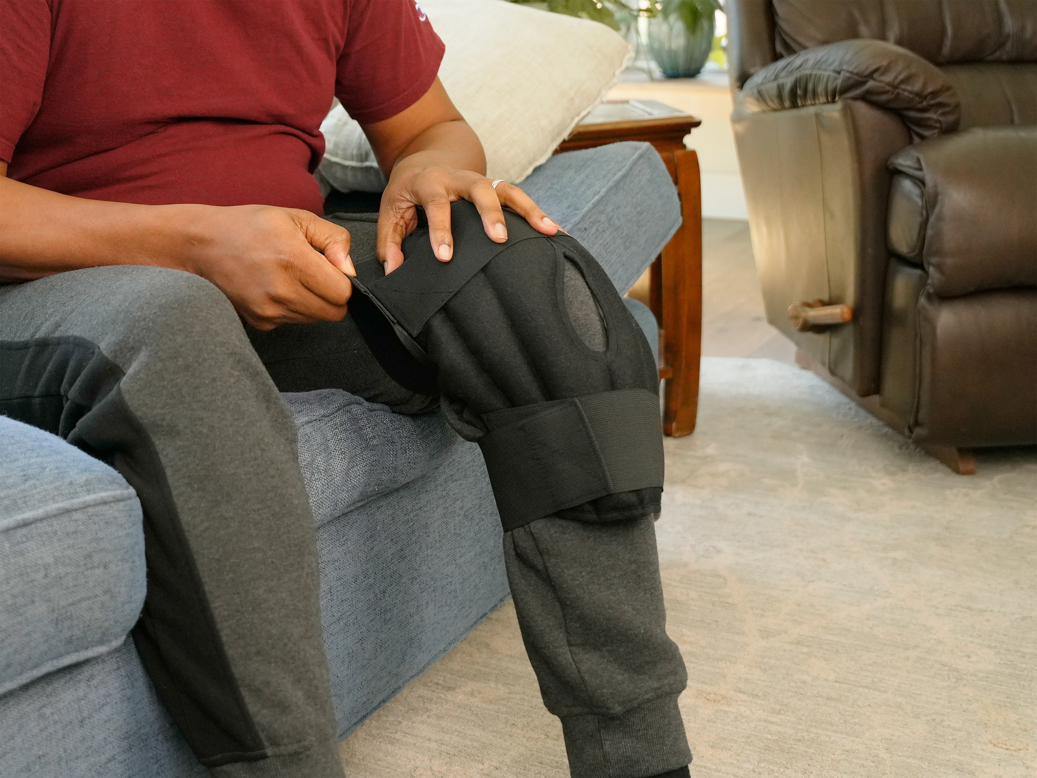 A person seated on a couch wearing a Moist Heat Thera-Temp Knee wrap with black straps over gray sweatpants in a living room setting.