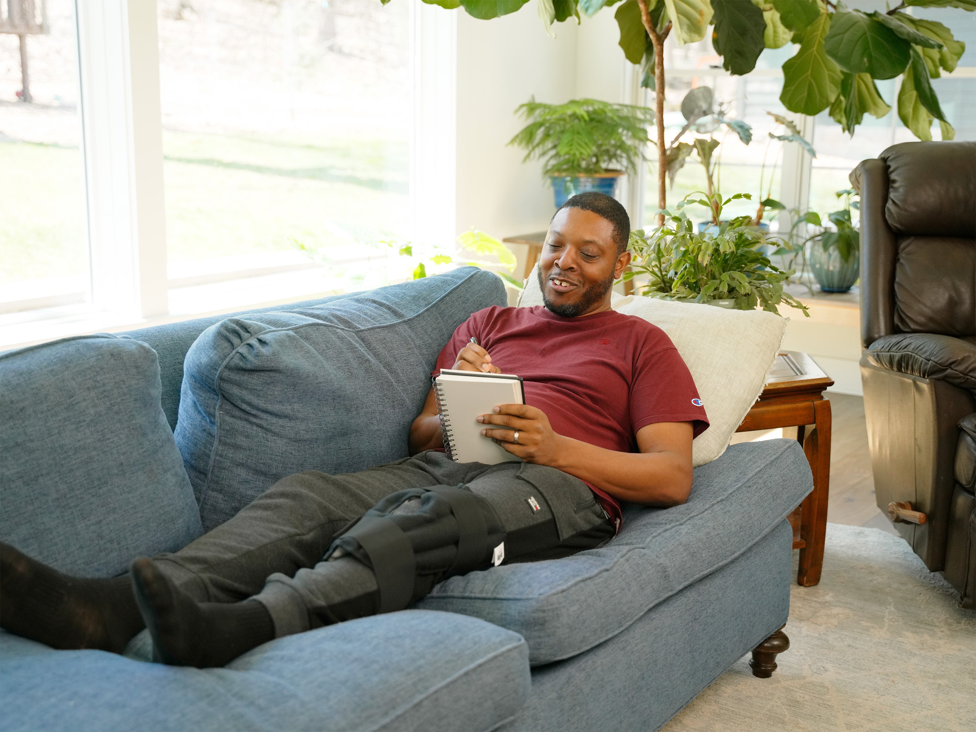A person seated on a couch wearing a Moist Heat Thera-Temp Knee wrap with black straps over gray sweatpants in a living room setting.