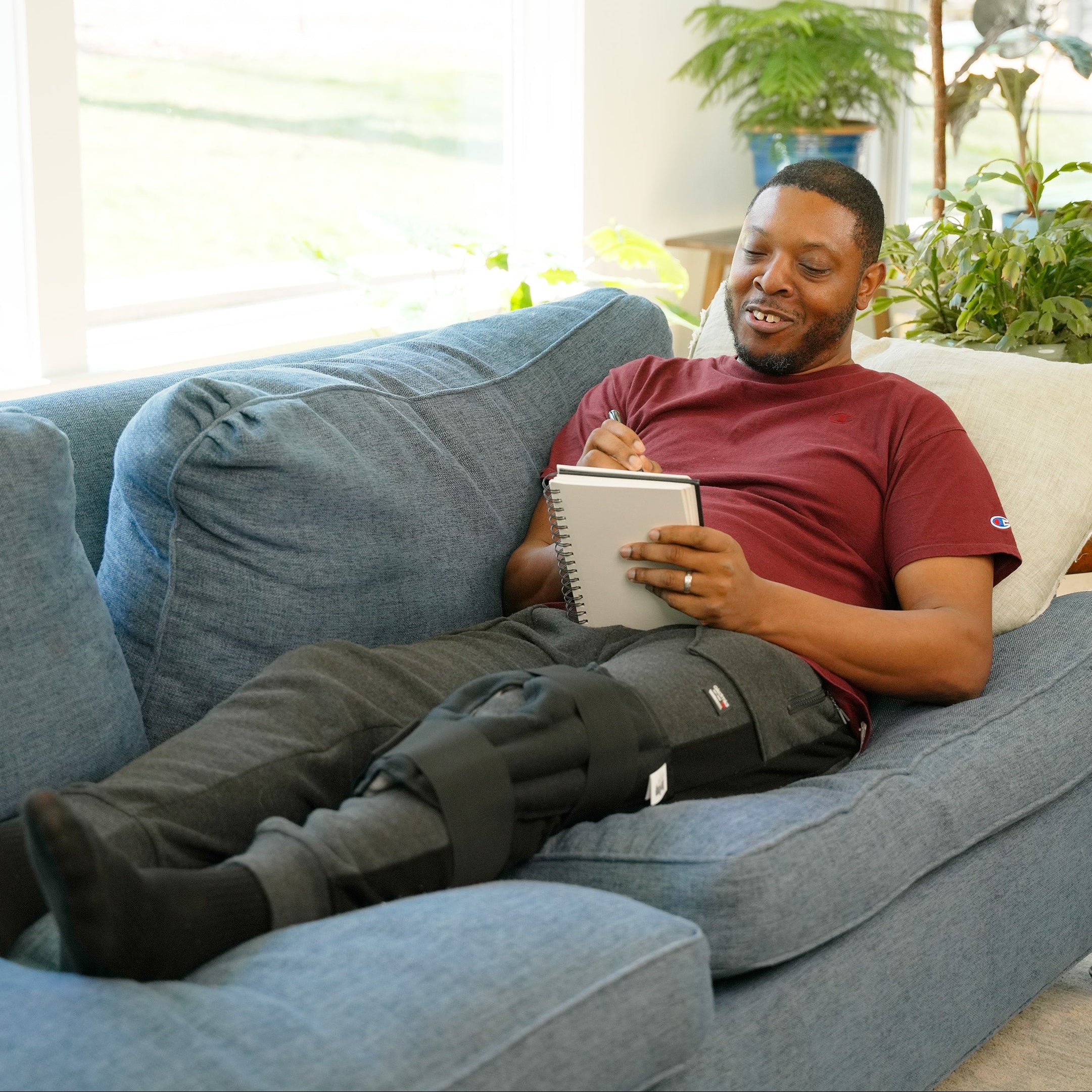 A man reclining on a blue couch in a bright living room, holding a tablet and wearing a Moist Heat Thera-Temp wrap on his knee.