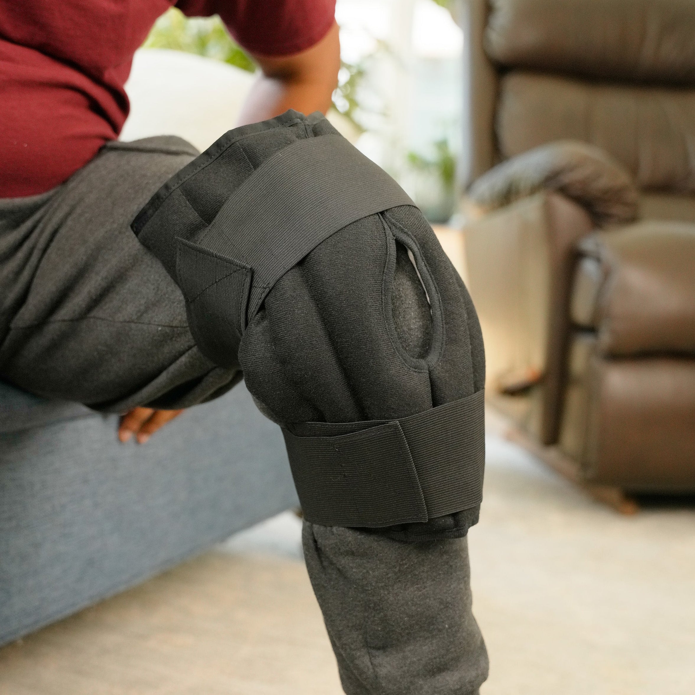 A person seated on a couch wearing a Moist Heat Thera-Temp Knee wrap with black straps over gray sweatpants in a living room setting.