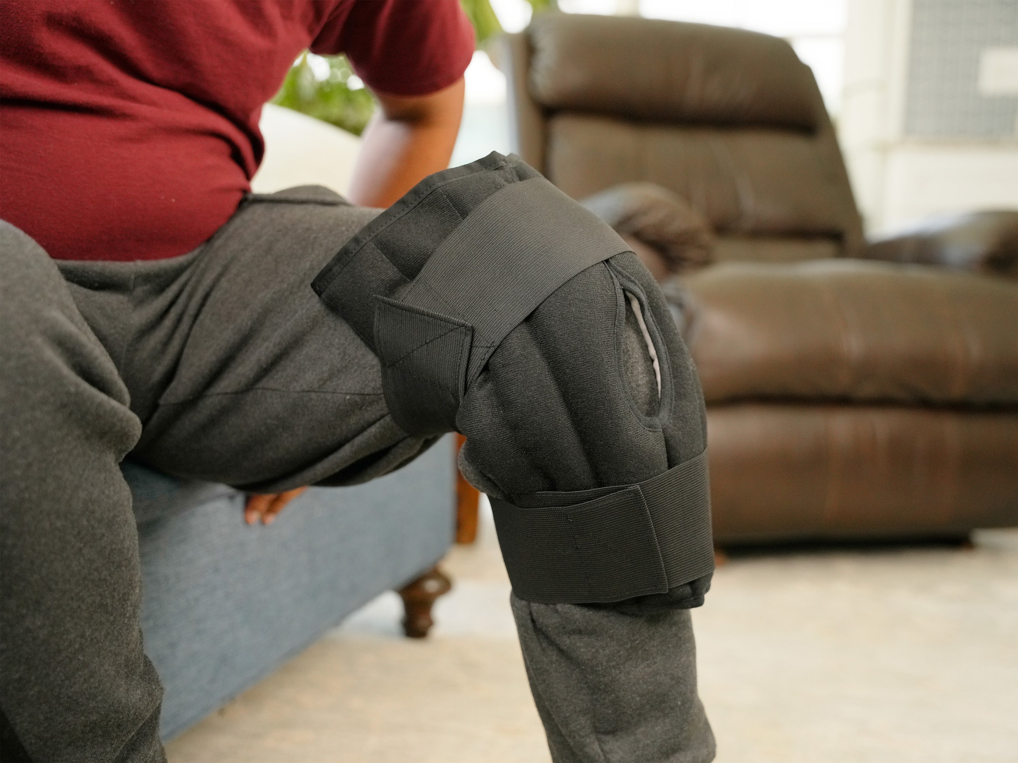 A person seated on a couch wearing a Moist Heat Thera-Temp Knee wrap with black straps over gray sweatpants in a living room setting.