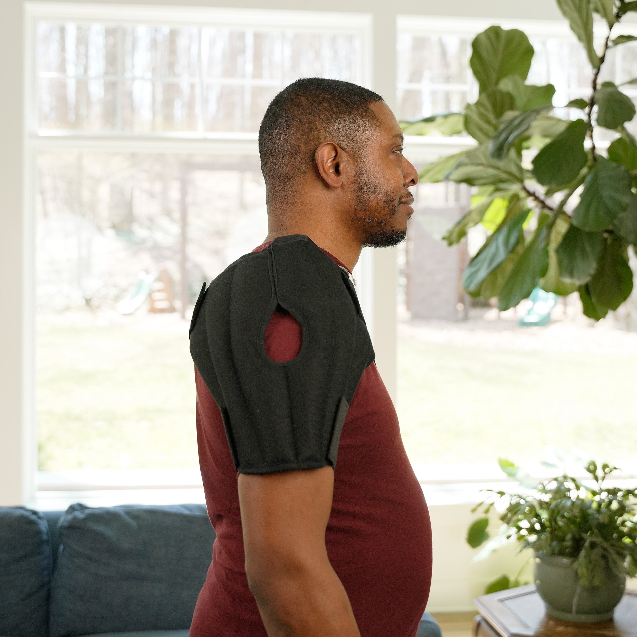 A man in profile wearing a black shoulder Moist Heat Thera-Temp wrap over a maroon t-shirt, standing in a brightly lit living room with a window, couch, and potted plant.
