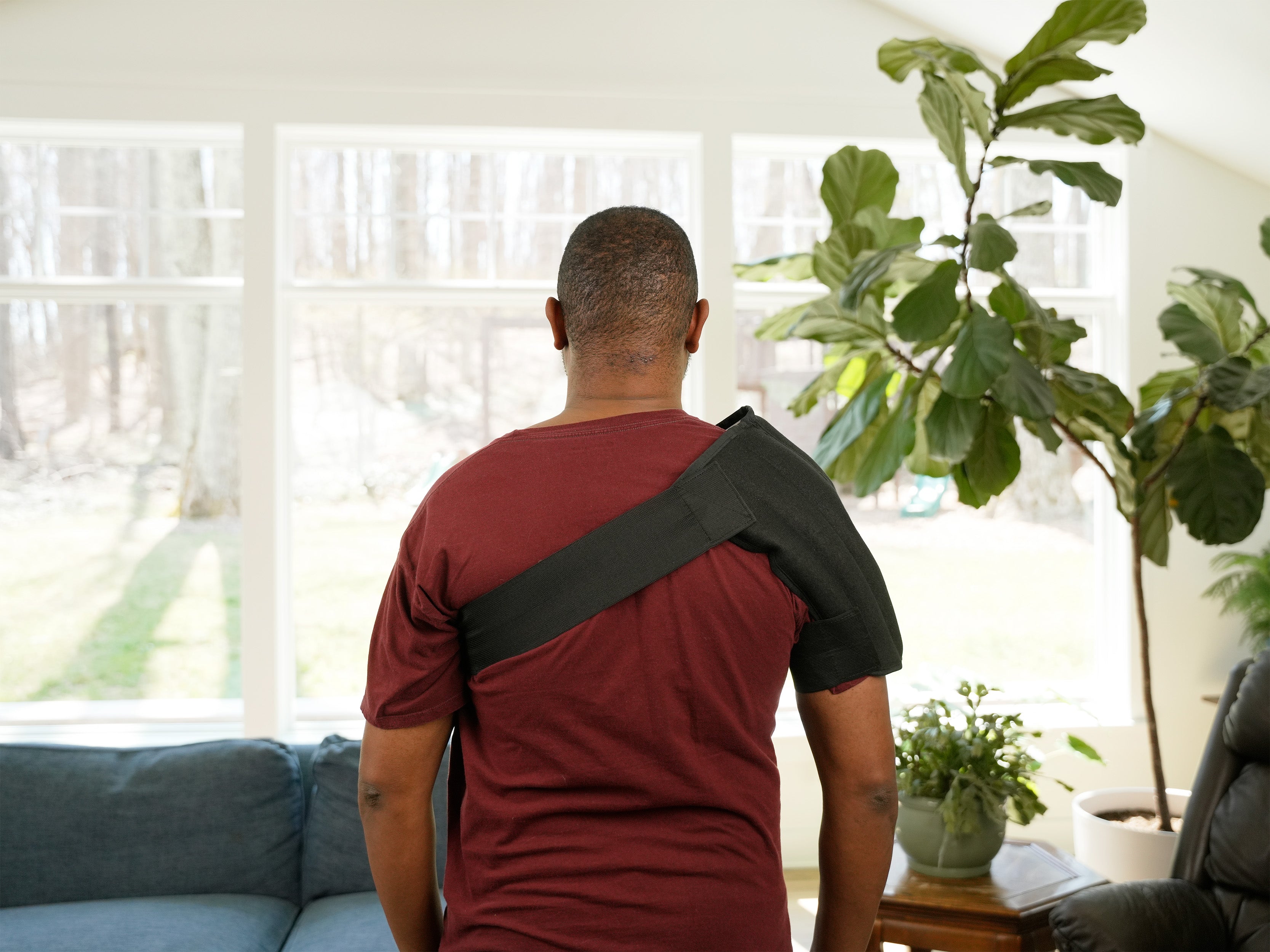 A man in profile wearing a black shoulder Moist Heat Thera-Temp wrap over a maroon t-shirt, standing in a brightly lit living room with a window, couch, and potted plant.