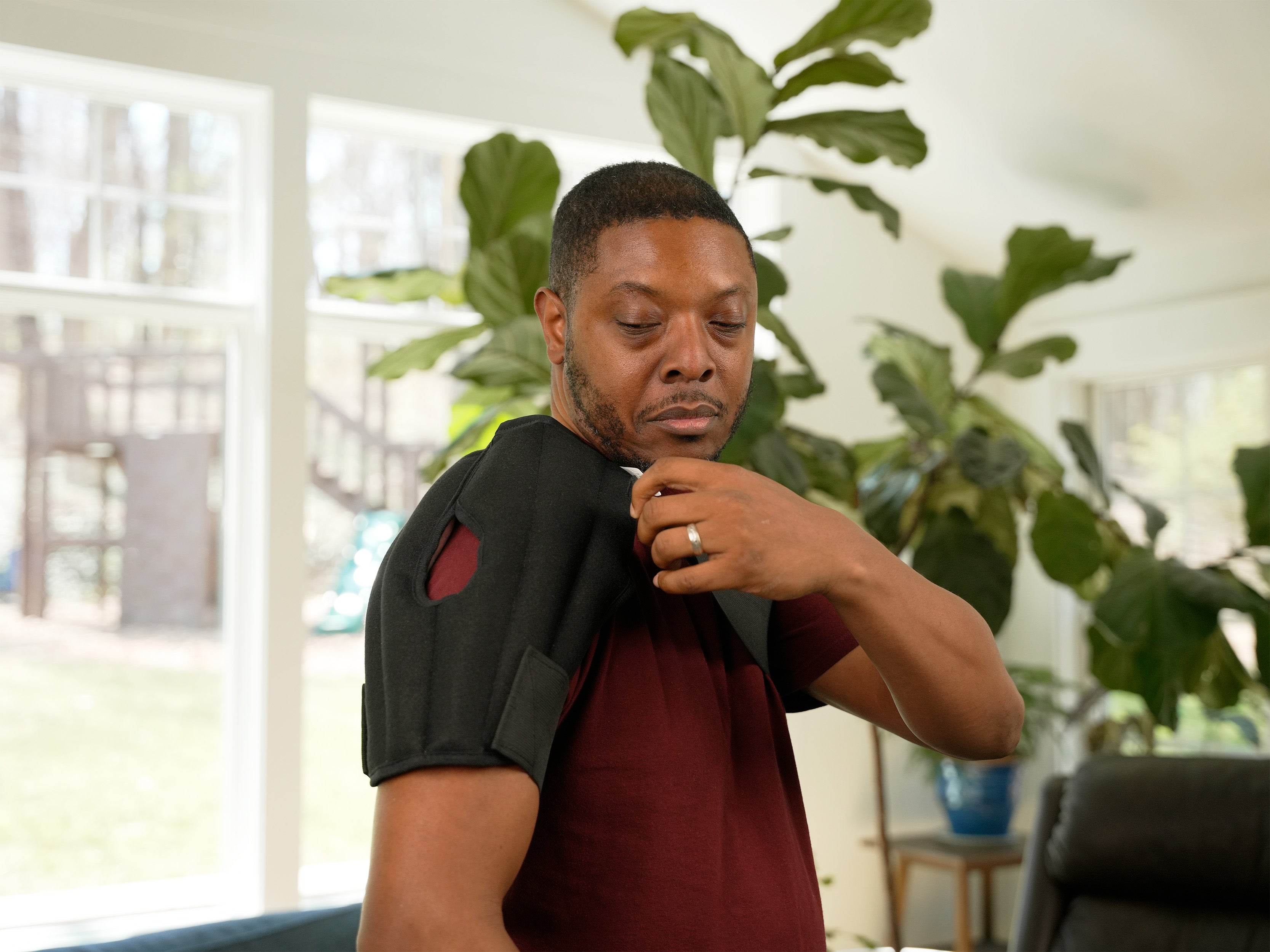A man in profile wearing a black shoulder Moist Heat Thera-Temp wrap over a maroon t-shirt, standing in a brightly lit living room with a window, couch, and potted plant.