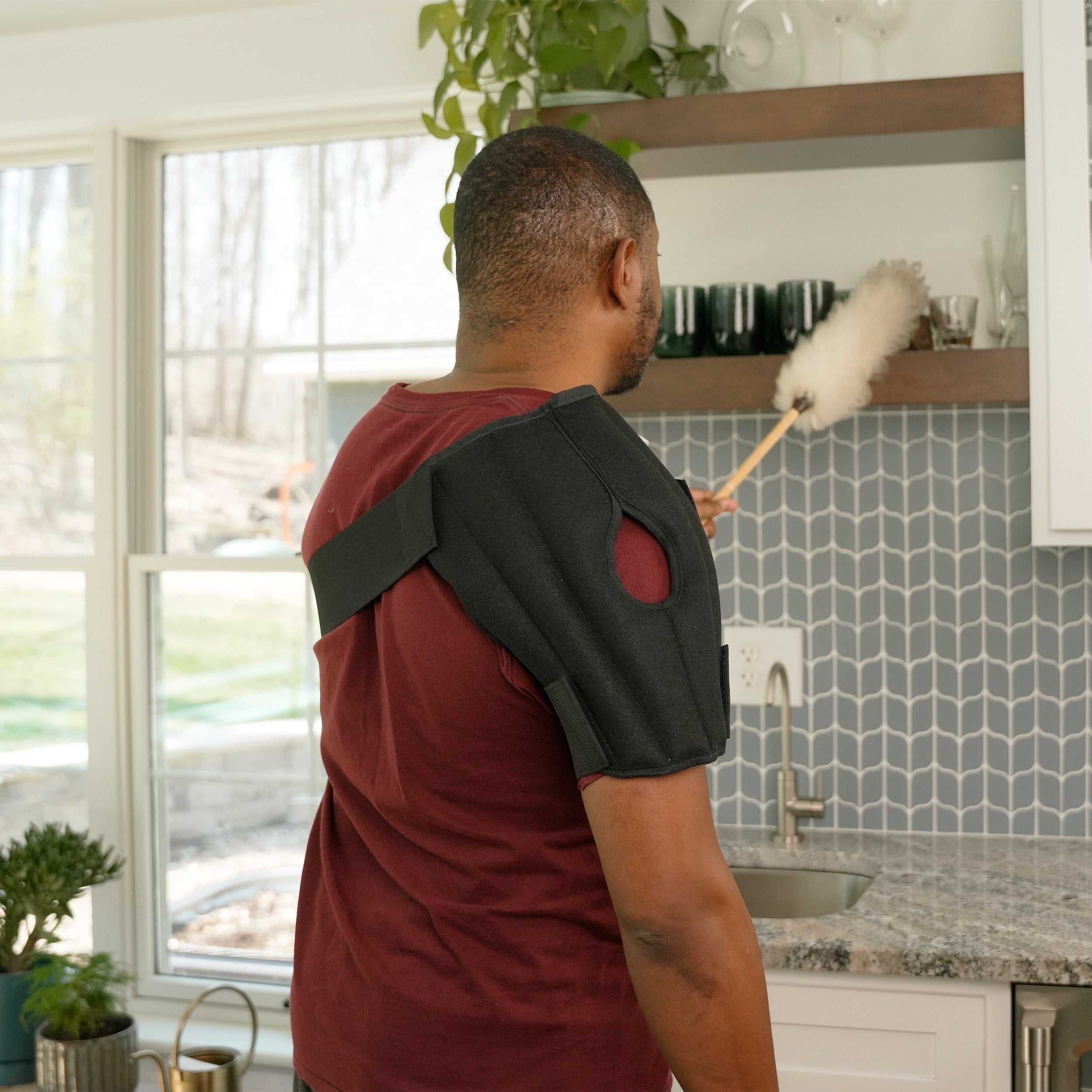A man in profile wearing a black shoulder Moist Heat Thera-Temp wrap over a maroon t-shirt, standing in a brightly lit wet bar dusting off the shelf.