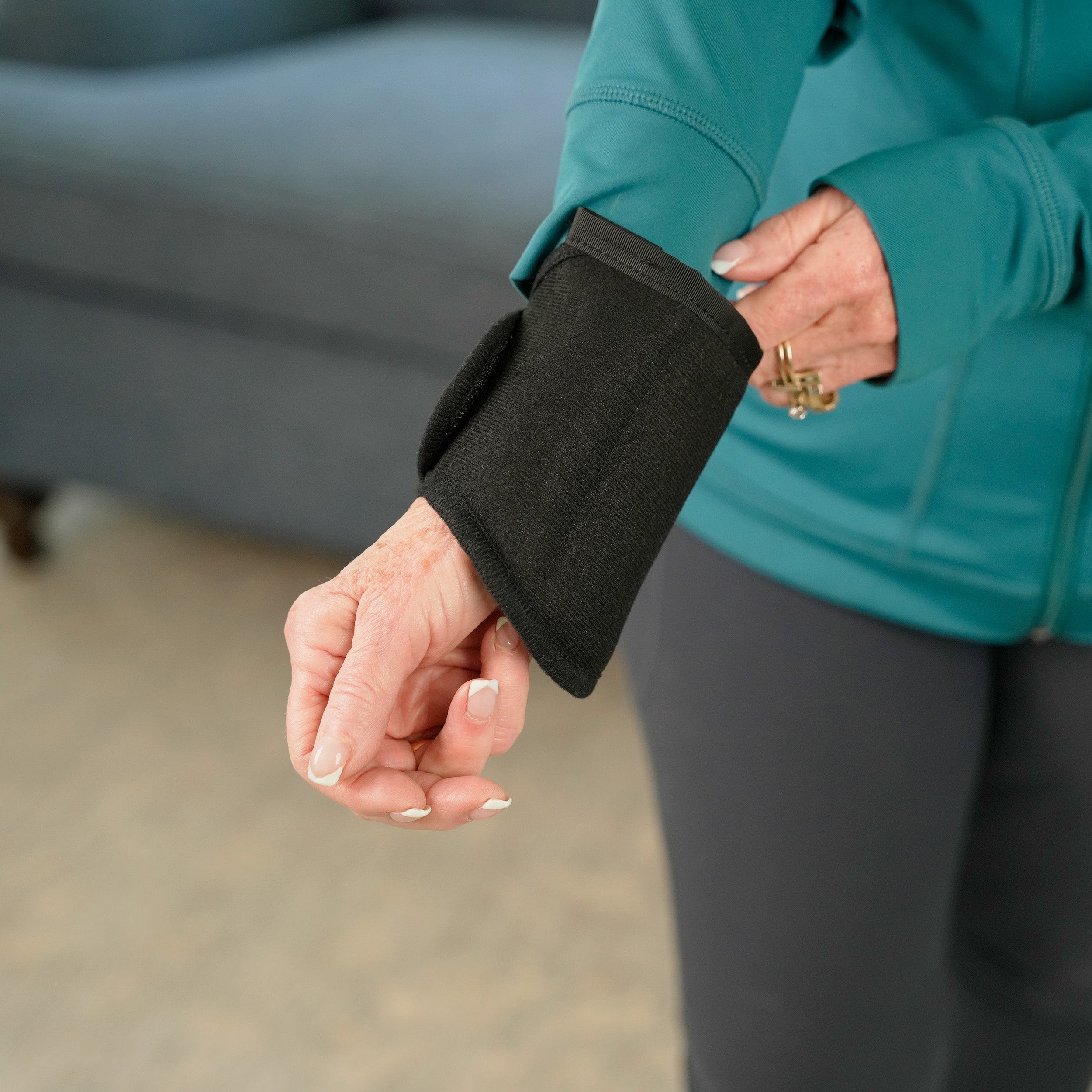 A close-up of a person wearing a black Moist Heat Thera-Temp Wrap on their forearm, with a teal zip-up jacket and dark leggings in a softly blurred indoor setting.