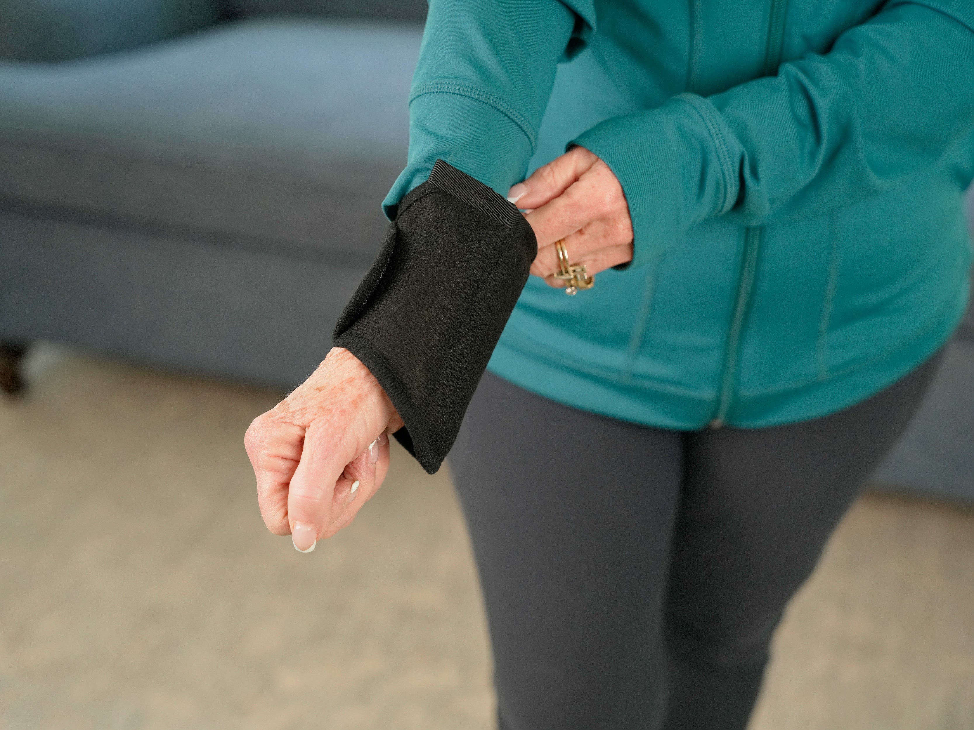 A close-up of a person wearing a black Moist Heat Thera-Temp Wrap on their forearm, with a teal zip-up jacket and dark leggings in a softly blurred indoor setting.