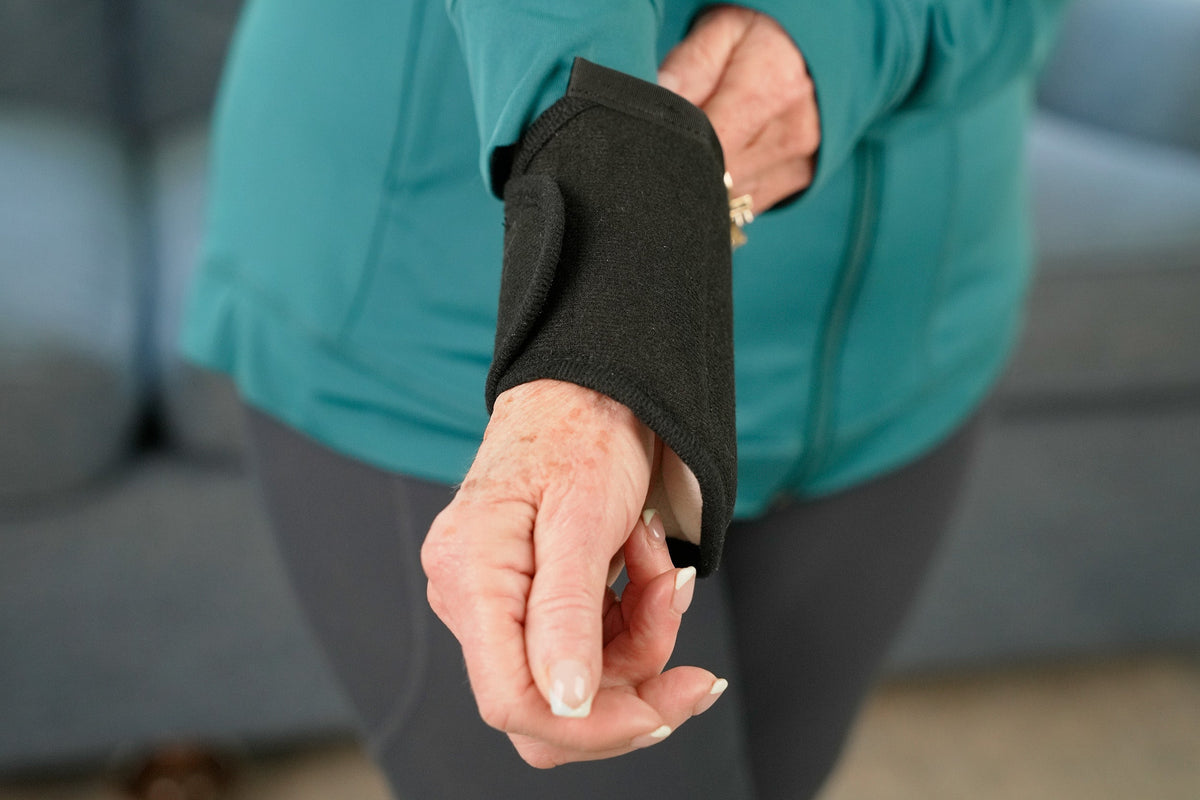 A close-up of a person wearing a black thera-temp moist heat wrist wrap on their left arm, with a teal zip-up jacket and gray leggings in the background.