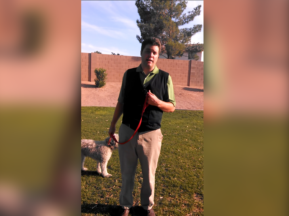A man standing outdoors on a grassy lawn holding a red leash next to a small white dog. He is wearing a black fashion cooling vest over a green polo shirt and khaki pants, with a brick wall and trees in the background.