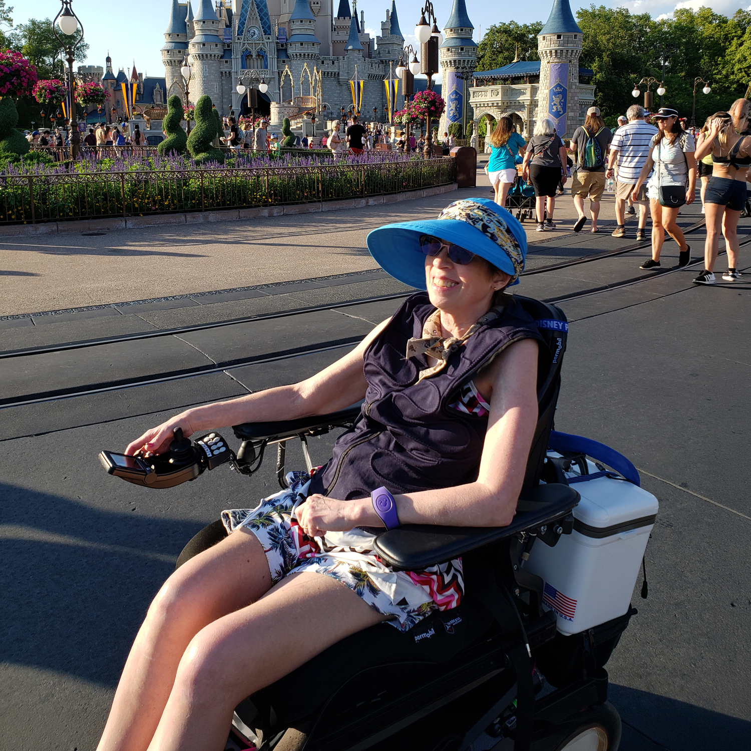 A woman in a motorized wheelchair wearing a blue sun hat and sunglasses sits on a pathway at a theme park, with a castle and other visitors in the background.