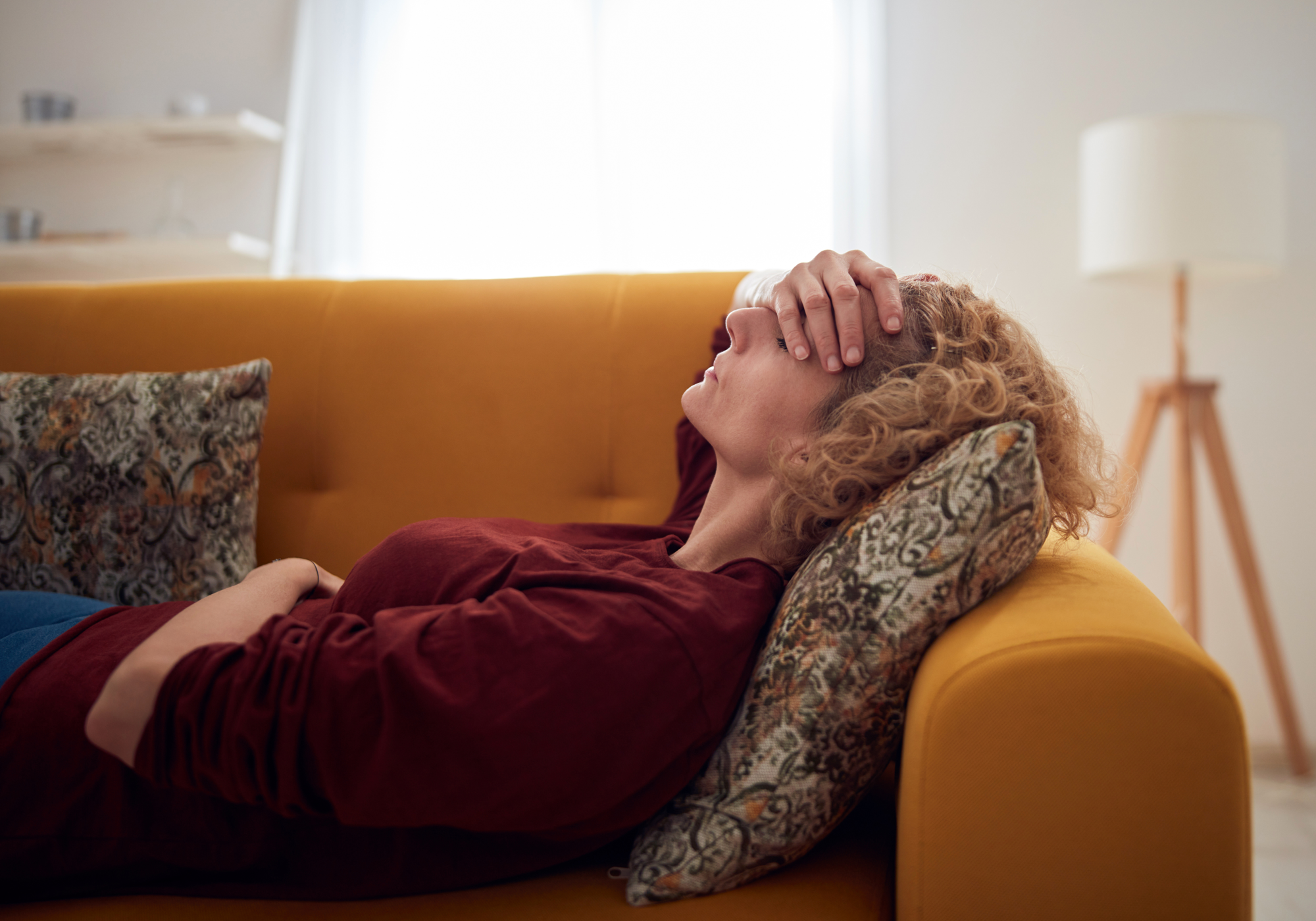 Woman lying on a yellow couch with a pillow, looking distressed.