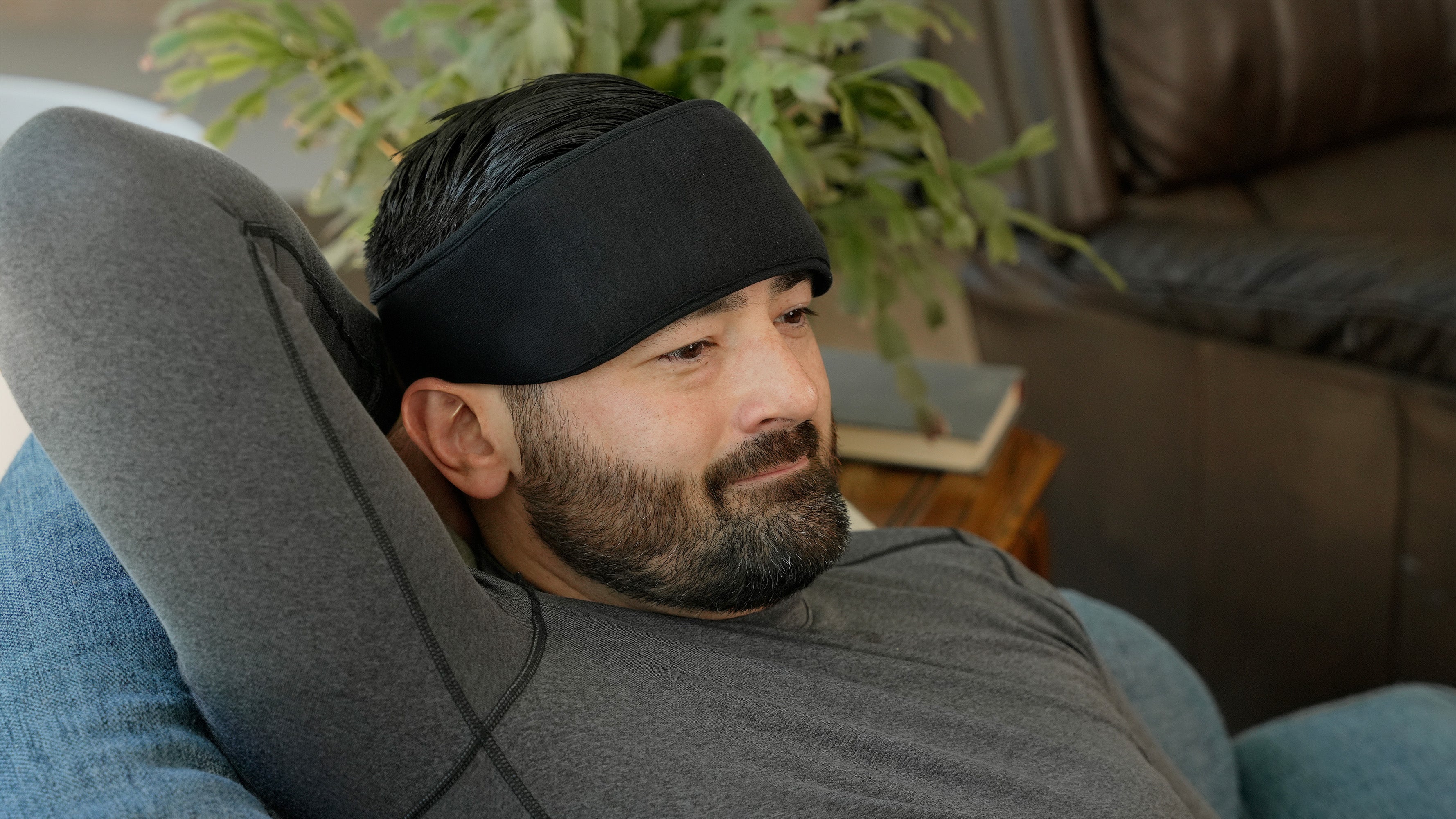 A man reclining on a gray lounge chair wearing a black Head and Neck Soft ice® mask and a gray long-sleeve shirt in a living room setting.