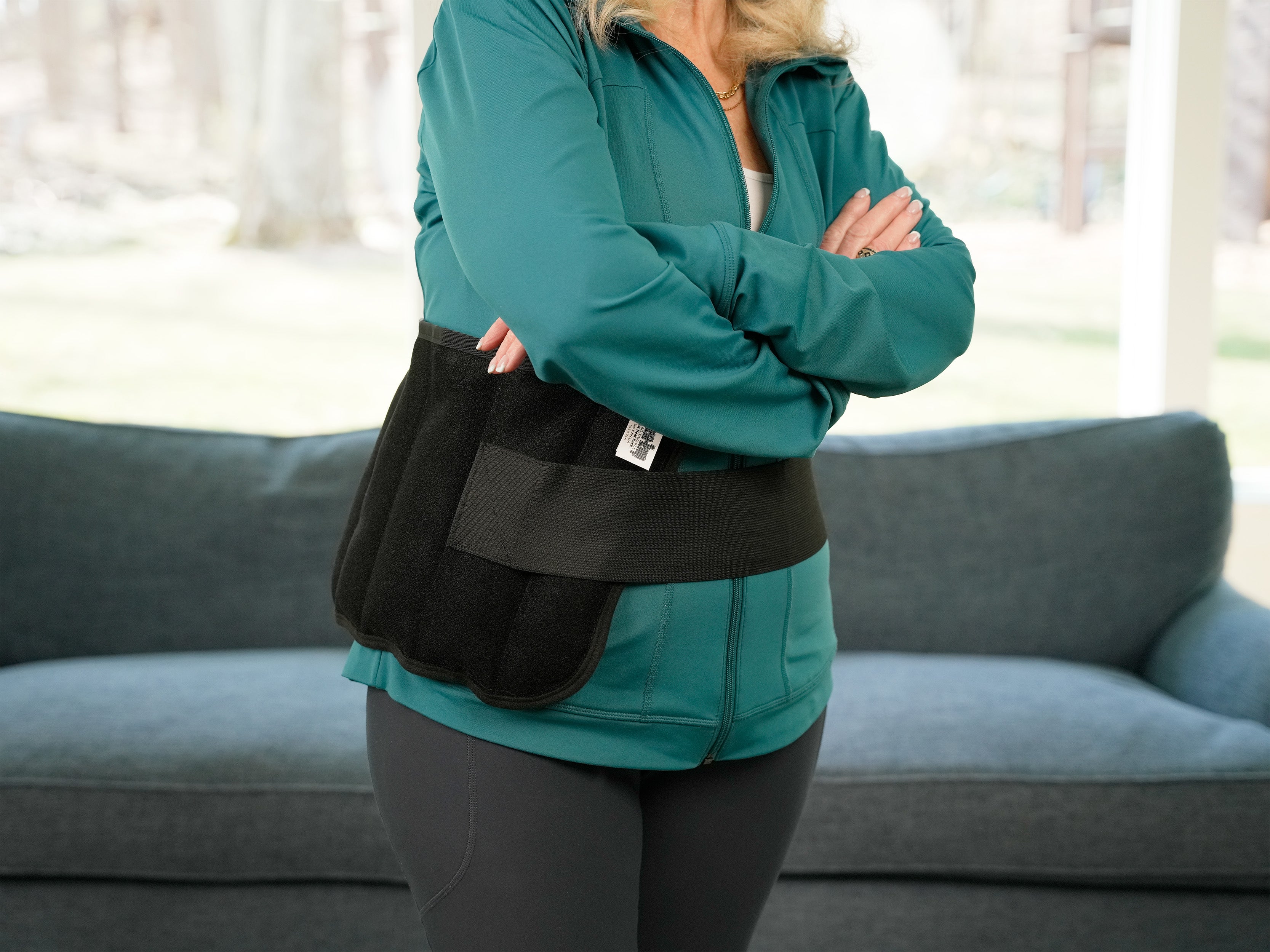 A woman wearing a teal zip-up jacket and dark leggings stands indoors with her arms crossed, wearing a black Moist Heat Thera-Temp wrapped around her lower torso. A blue-gray couch is visible in the background.